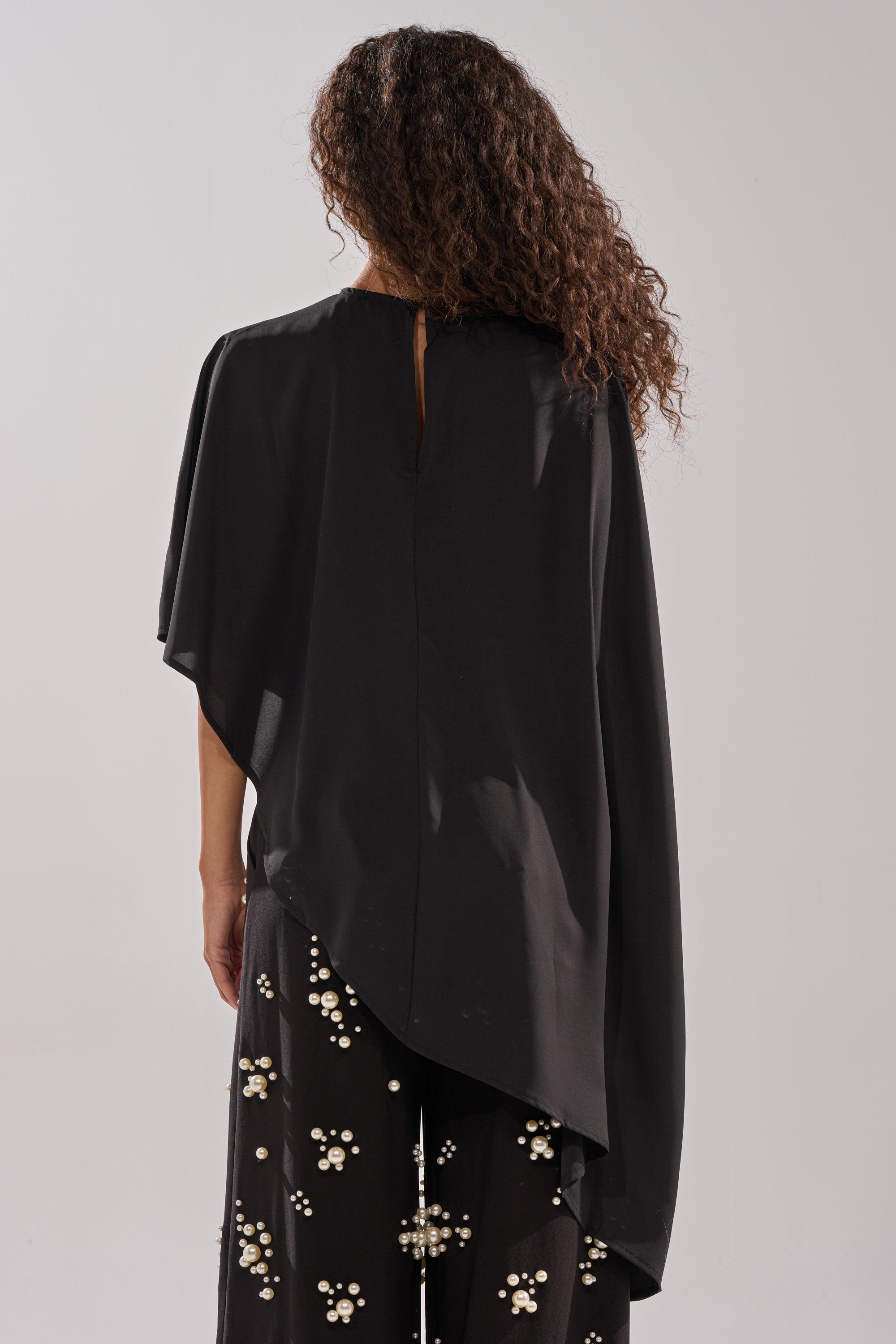 A person with long, curly hair is shown from the back wearing the SOMETHING DIFFERENT OVERSIZED ASYMMETRICAL TEE, styled with black pants featuring white and gold floral patterns.