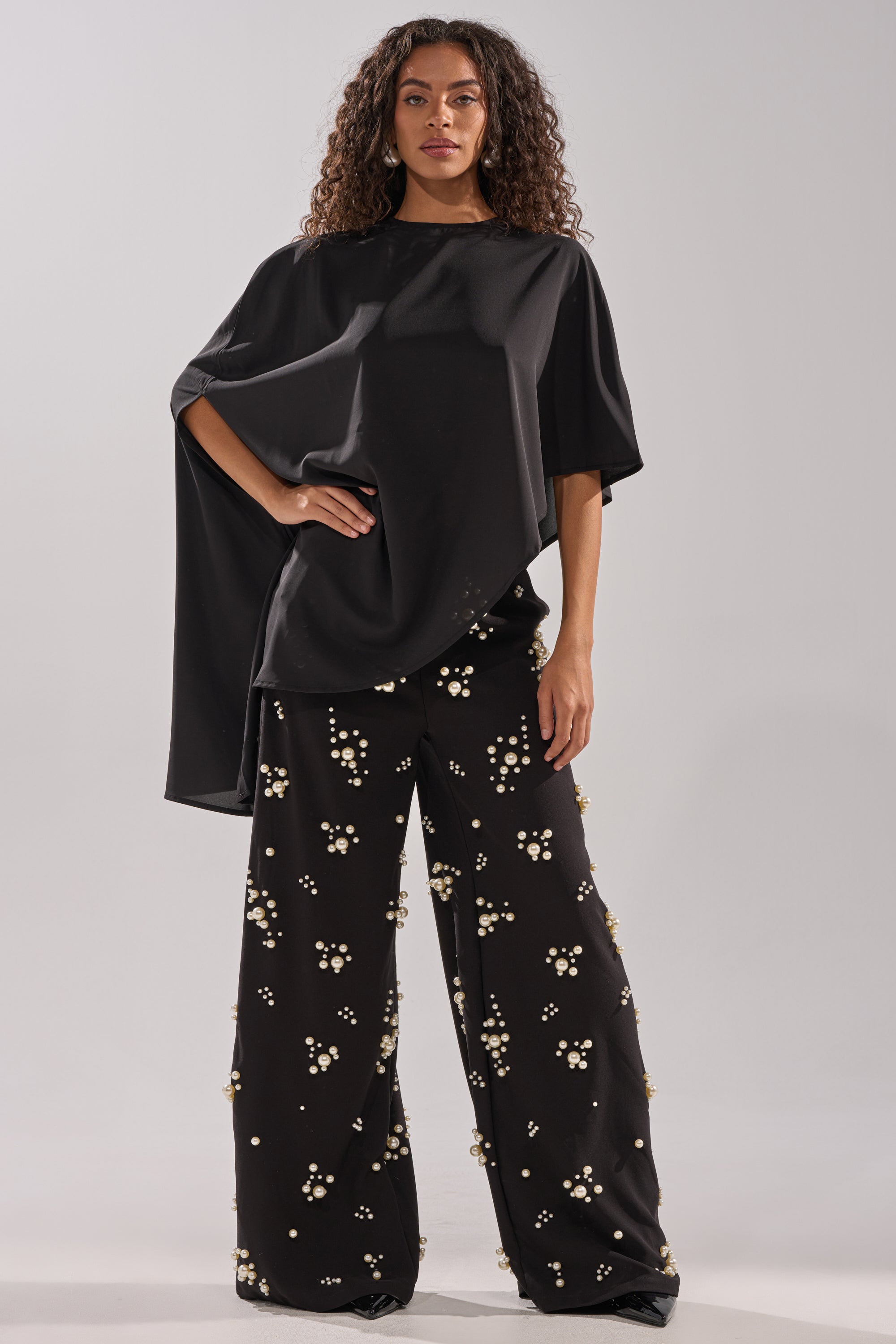 A woman with curly hair wears the SOMETHING DIFFERENT OVERSIZED ASYMMETRICAL TEE and wide-leg black pants embellished with pearls and beads, standing against a plain light background.
