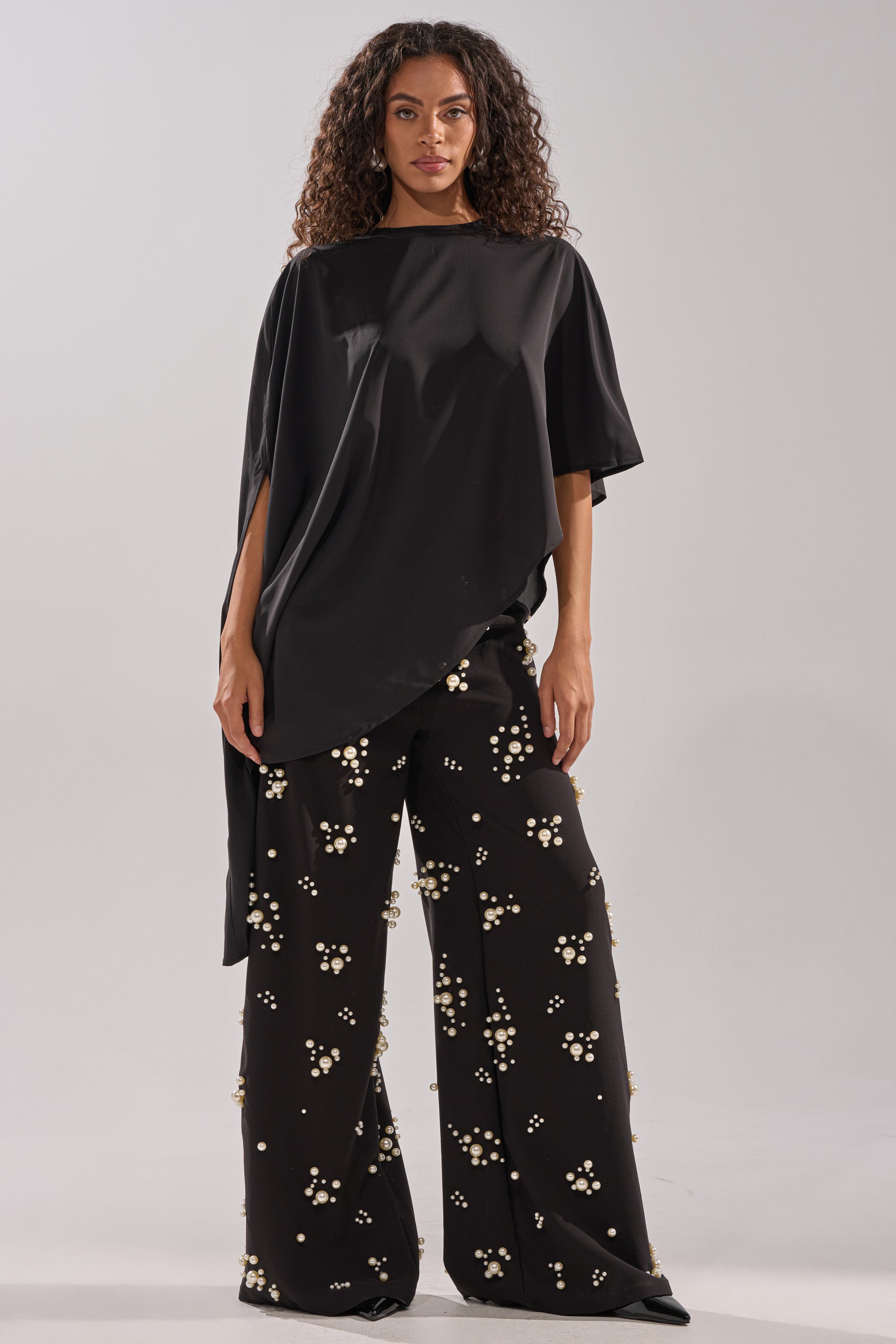 A woman with curly hair wears the SOMETHING DIFFERENT OVERSIZED ASYMMETRICAL TEE and wide-leg black pants with white beaded designs, standing against a plain light gray background.