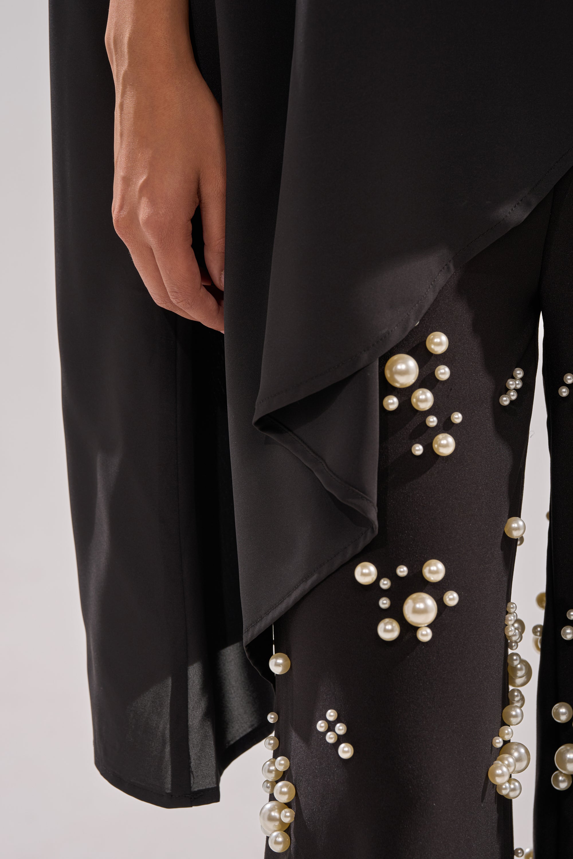 A close-up of a person wearing black pants with pearl-like beads and the SOMETHING DIFFERENT OVERSIZED ASYMMETRICAL TEE, their hand resting by the layered hem to highlight the cape-like silhouette of this statement top.