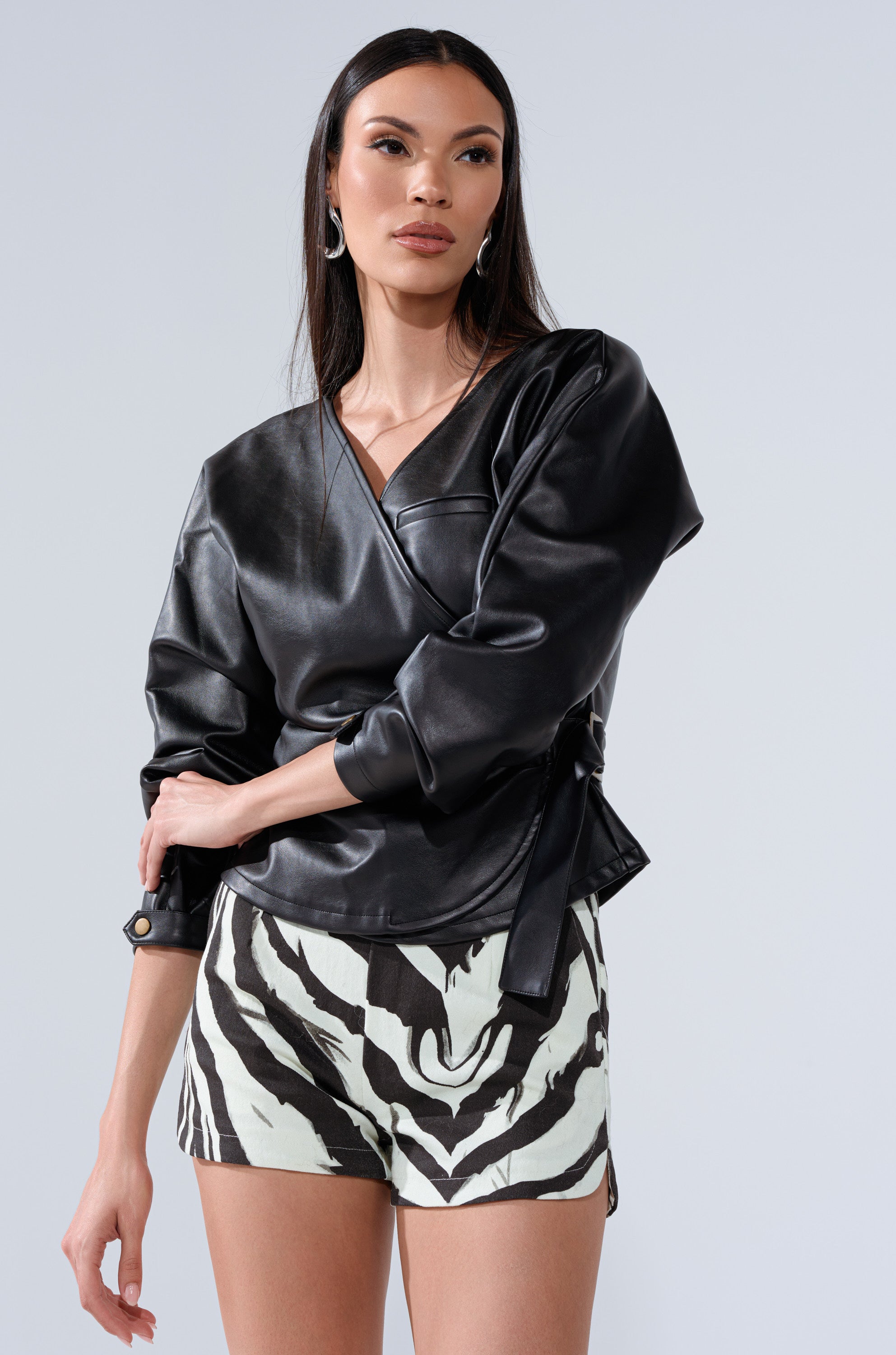 A woman poses in the EASY ENOUGH FAUX LEATHER TIE FRONT BLOUSE, styled as a wrap jacket, paired with high-waisted black and white zebra print shorts against a plain light background.