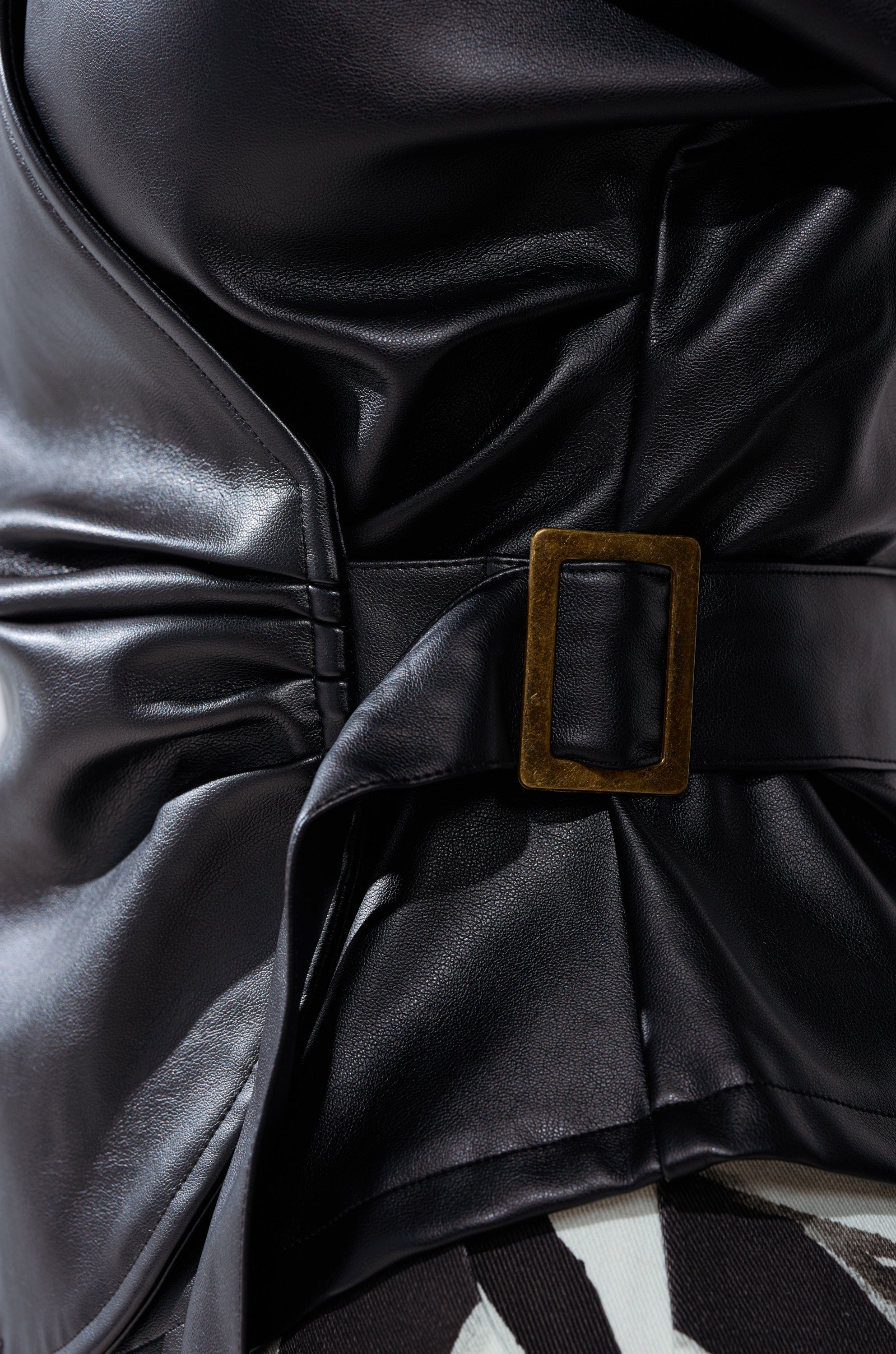 Close-up of the EASY ENOUGH FAUX LEATHER TIE FRONT BLOUSE featuring shiny vegan leather, a dark brass rectangular buckle, and gathered fabric that reveals a glimpse of the patterned black-and-white blouse underneath.
