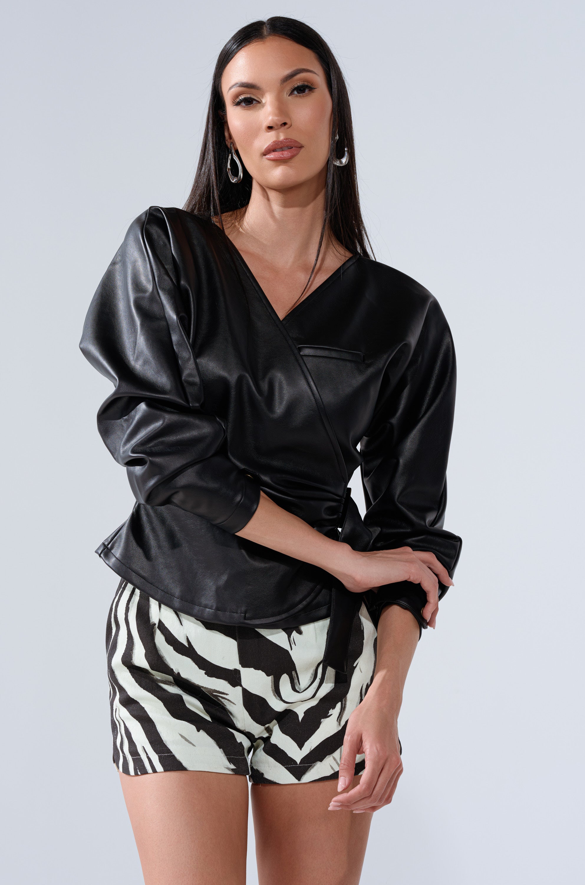 A woman with long dark hair poses against a plain light background wearing the EASY ENOUGH FAUX LEATHER TIE FRONT BLOUSE and black-and-white zebra print shorts, with one arm bent and the other relaxed.