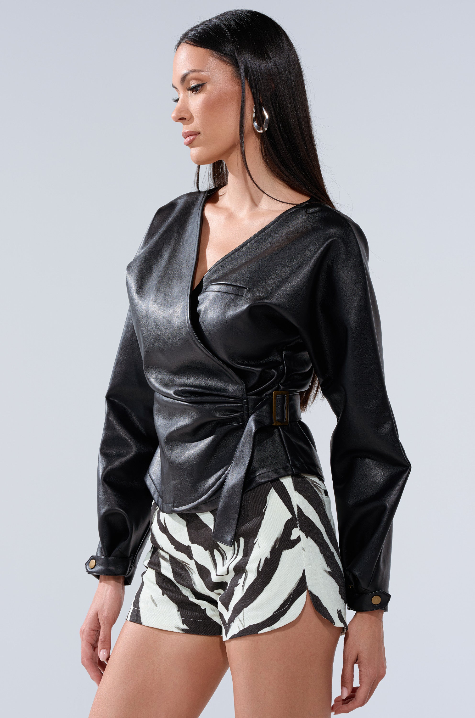 A woman with long dark hair faces sideways with a neutral expression, wearing the EASY ENOUGH FAUX LEATHER TIE FRONT BLOUSE and black-and-white zebra print shorts against a plain light gray background.