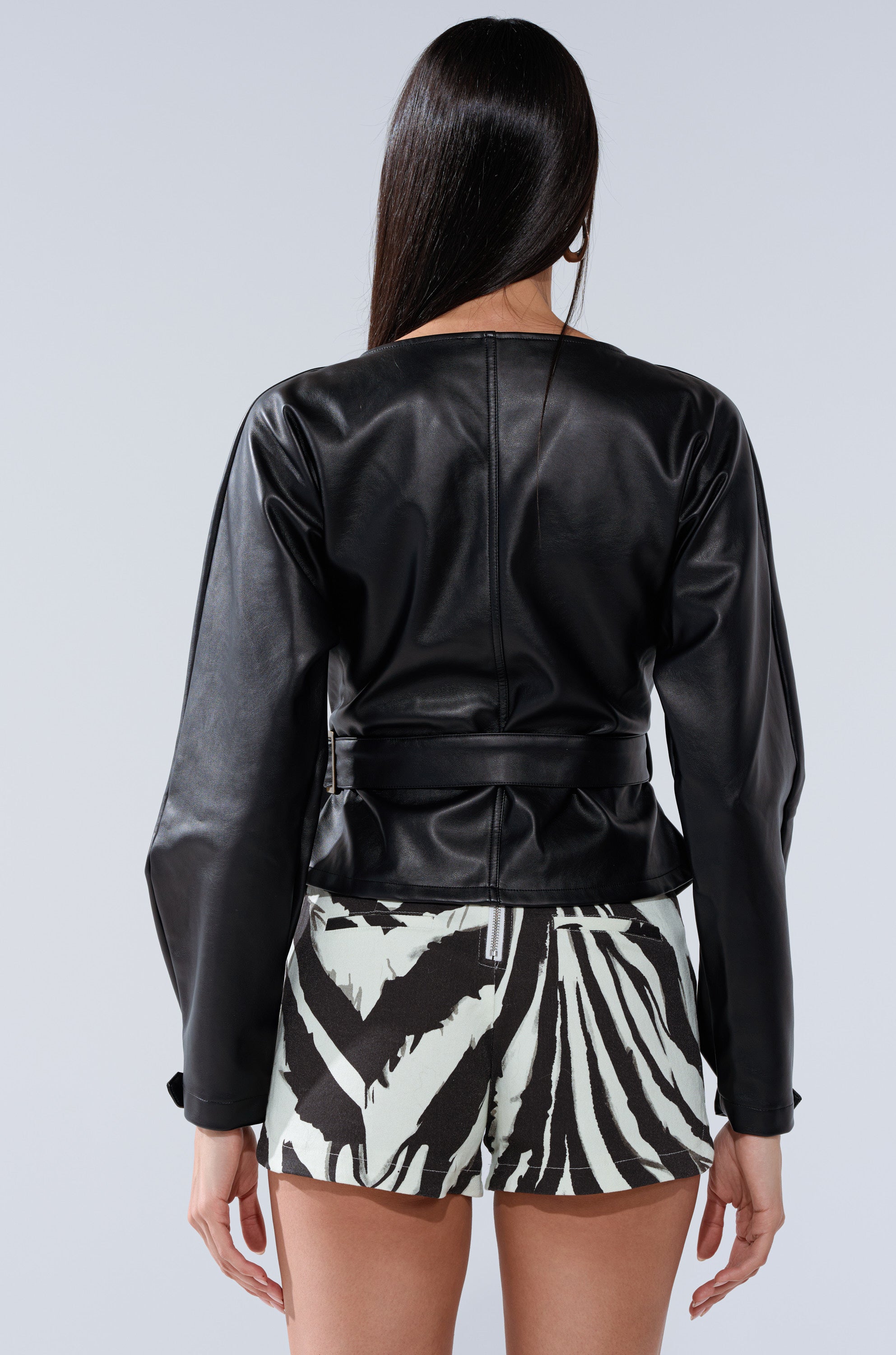 A woman with long dark hair stands with her back to the camera, wearing the EASY ENOUGH FAUX LEATHER TIE FRONT BLOUSE and black-and-white zebra print shorts.