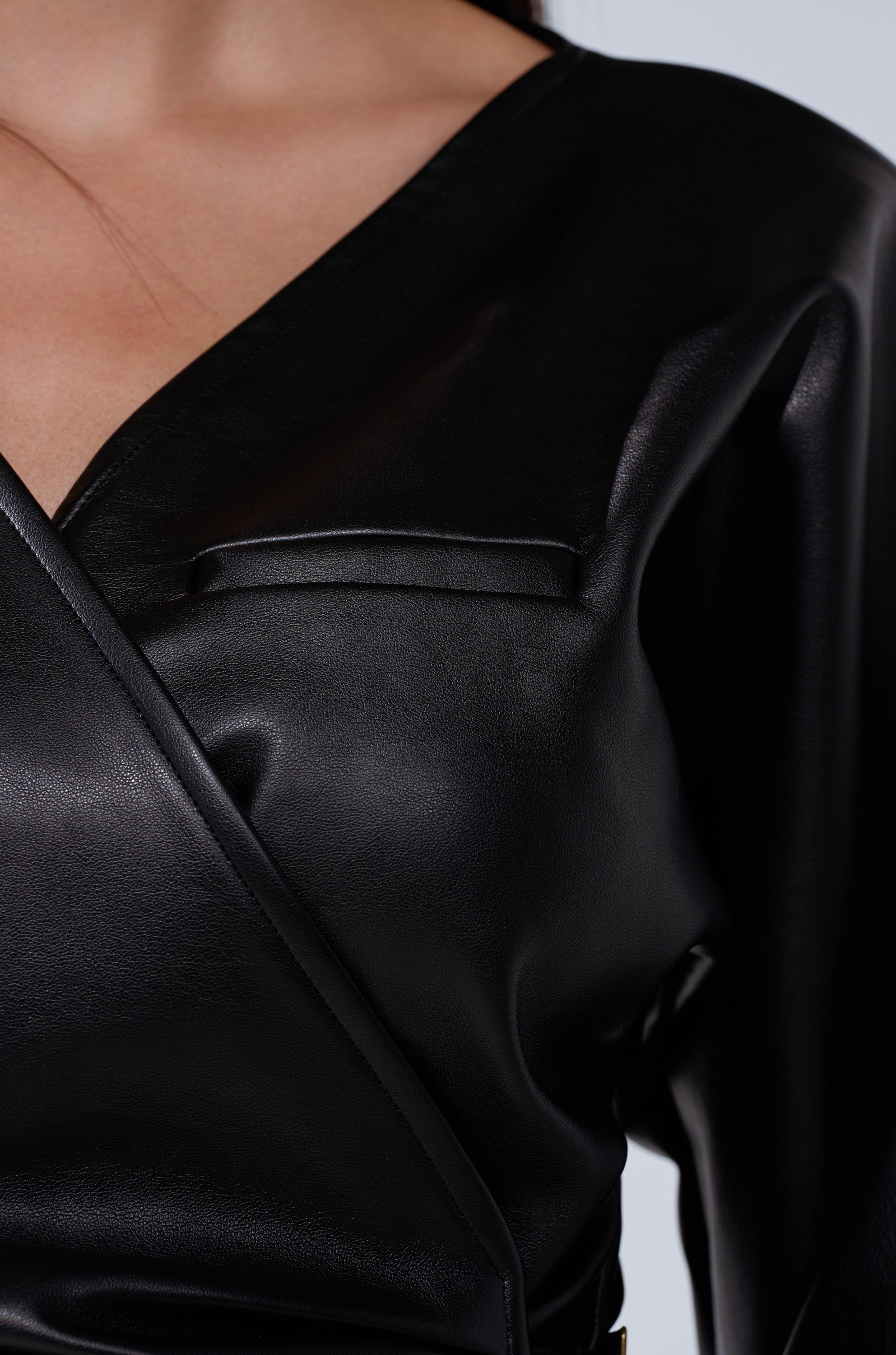 A close-up of someone wearing the EASY ENOUGH FAUX LEATHER TIE FRONT BLOUSE in black, featuring a V-neckline and chest pocket detail, highlighting the upper chest and shoulder.