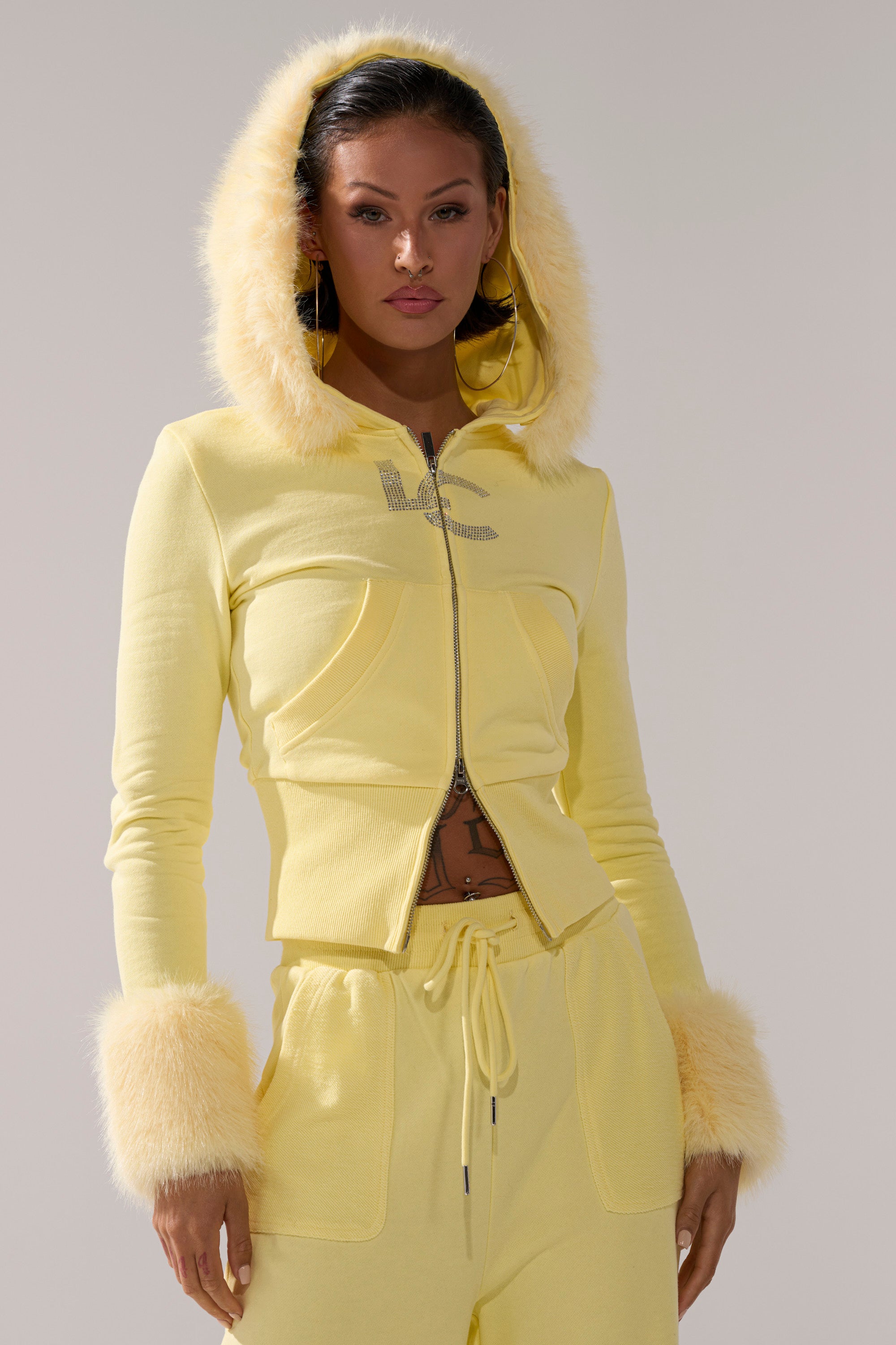 A woman wears the BUTTERFINGER ZIP UP SWEATSHIRT, a fitted light yellow hoodie with faux fur trim, rhinestone chest detail, furry hood and cuffs, paired with matching drawstring pants and a midriff-baring top.