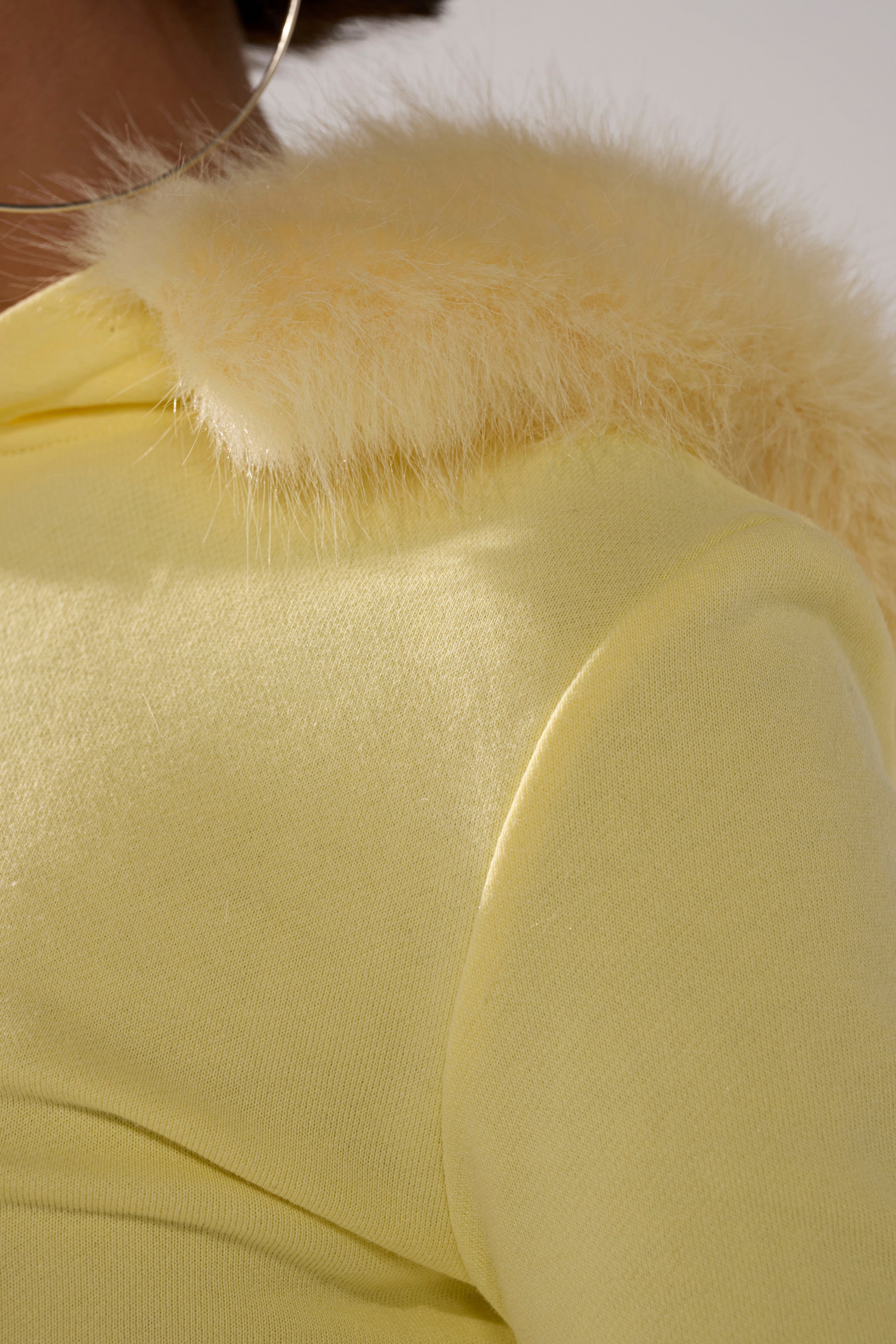 Close-up of a person in the BUTTERFINGER ZIP UP SWEATSHIRT, showcasing its pale yellow hue and faux fur collar trim, paired with a large hoop earring. The image emphasizes the soft texture and color of both clothing and accessory.