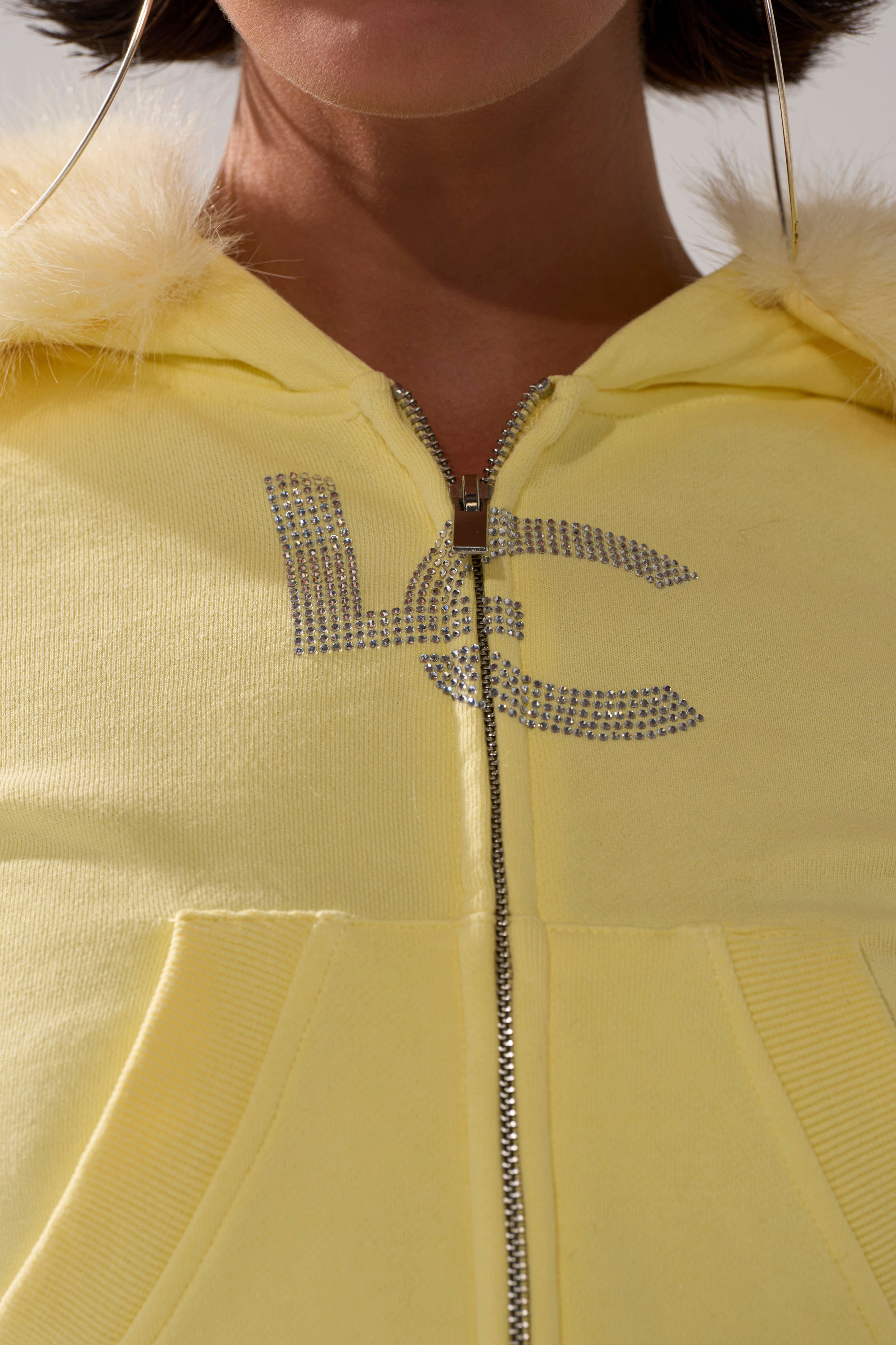 Close-up of a person wearing the BUTTERFINGER ZIP UP SWEATSHIRT with faux fur hood trim, rhinestone "LC" letters on the chest, visible pockets, and large hoop earrings for a cozy look.