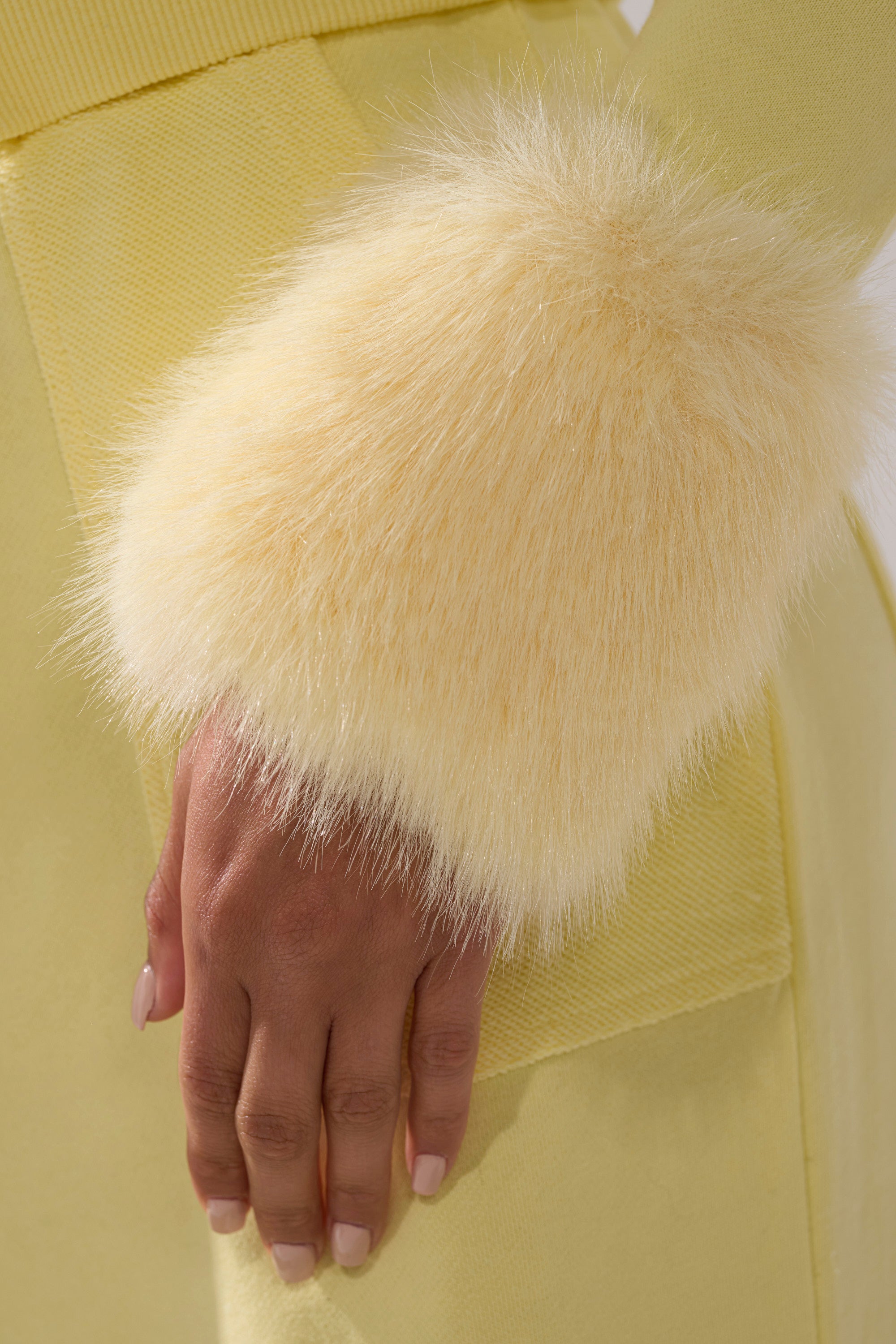 A close-up of a hand with a natural manicure resting on the BUTTERFINGER ZIP UP SWEATSHIRT, highlighting its fluffy, pale yellow faux fur trim cuff.