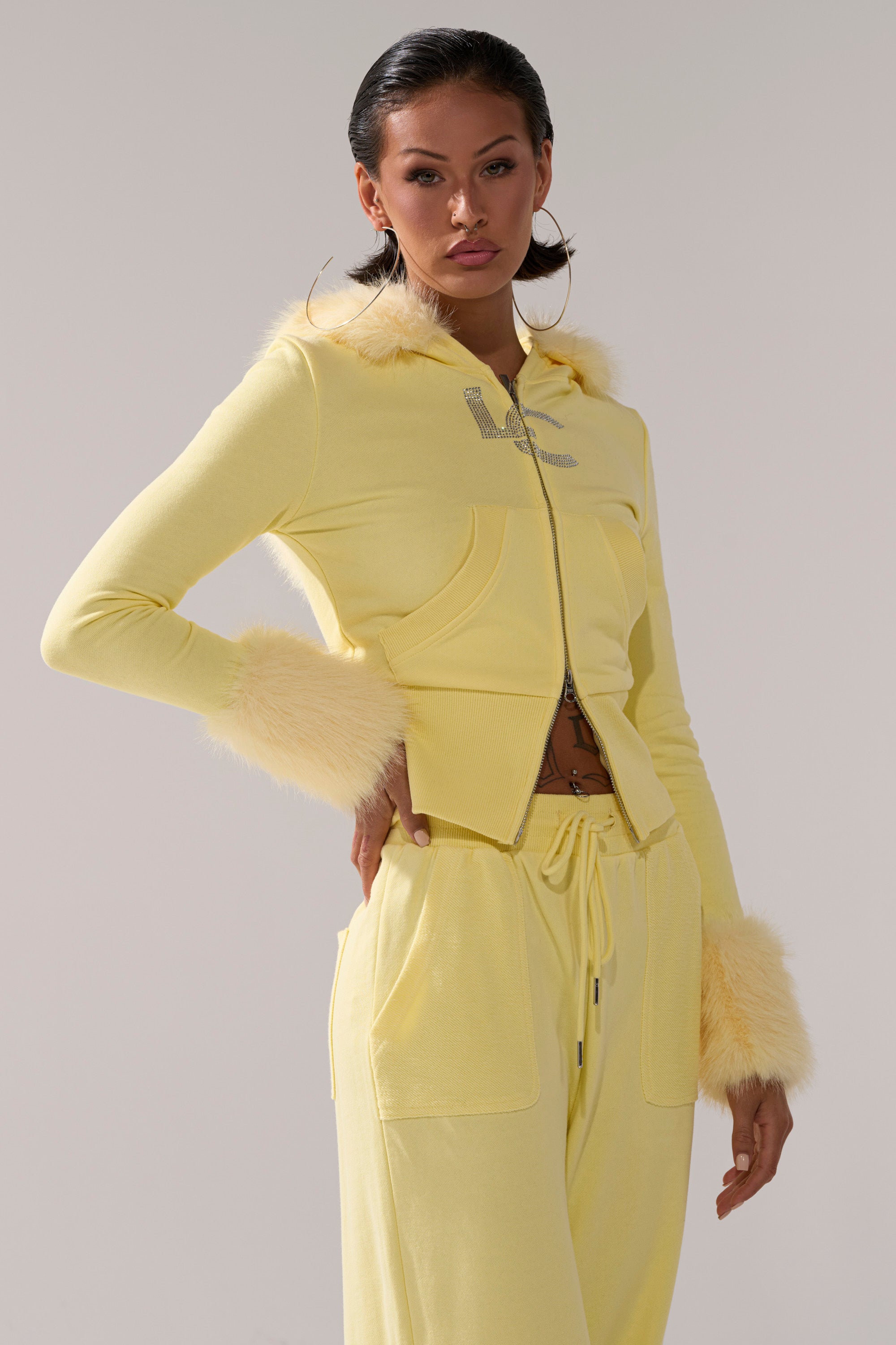 A woman poses with one hand on her hip against a light gray background, wearing the BUTTERFINGER ZIP UP SWEATSHIRT with matching faux fur-trimmed pants.