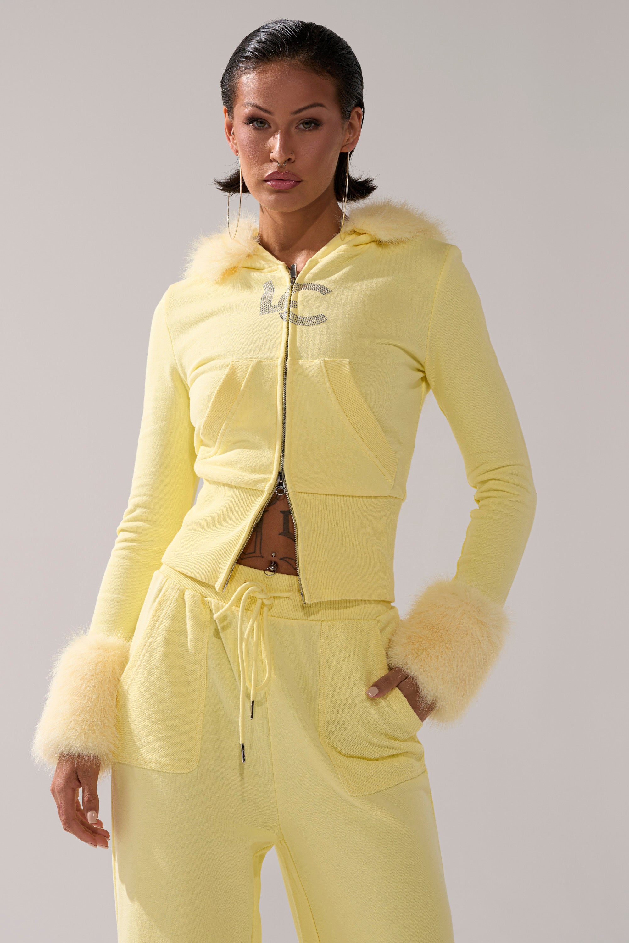 A person models the BUTTERFINGER ZIP UP SWEATSHIRT in light yellow faux fur trim, paired with matching drawstring pants, striking a confident pose with one hand in their pocket.