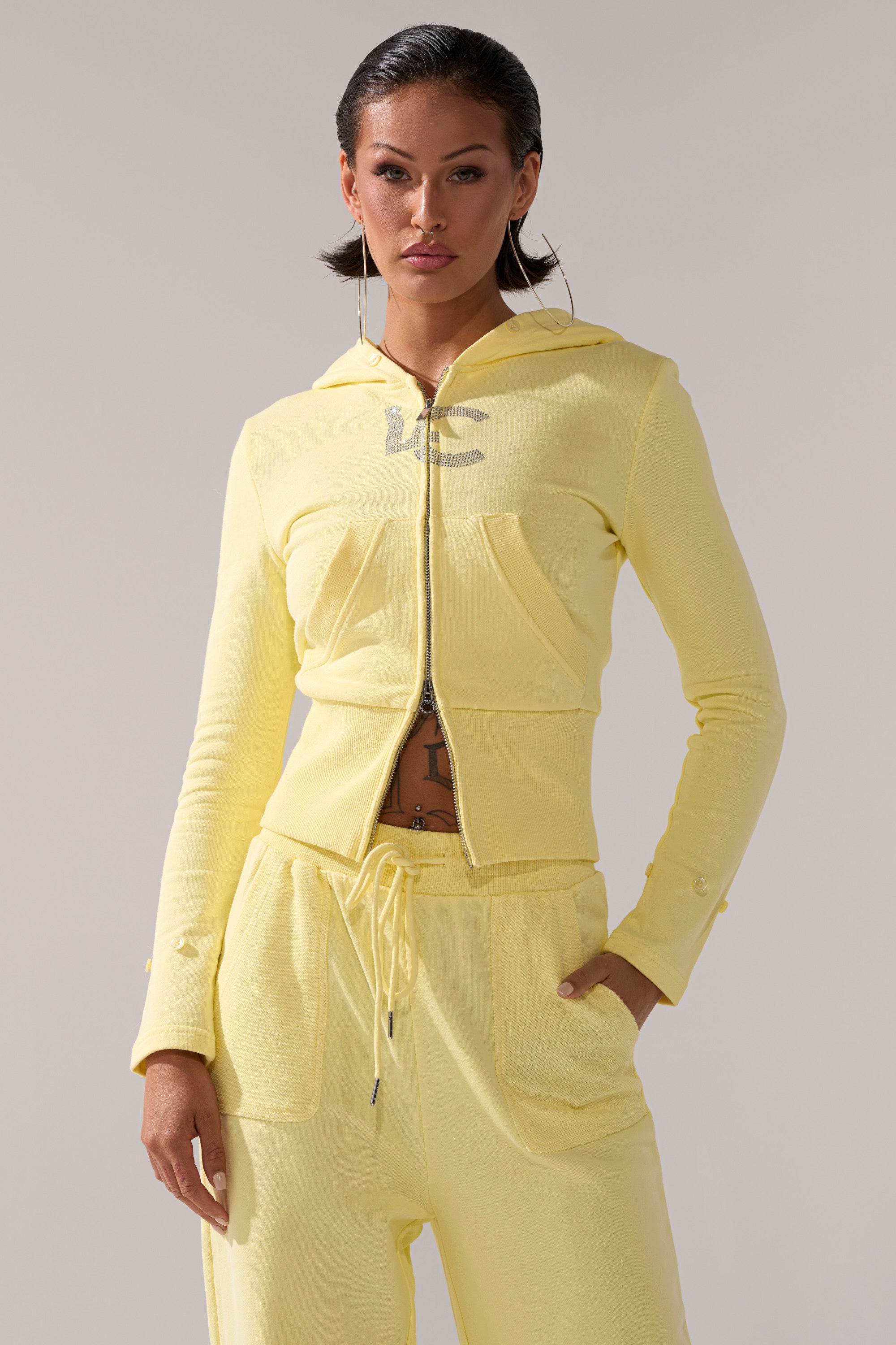 A woman stands confidently in a fitted, light yellow BUTTERFINGER ZIP UP SWEATSHIRT and matching drawstring pants. With her hair slicked back and large hoop earrings, she completes a bold, stylish look.
