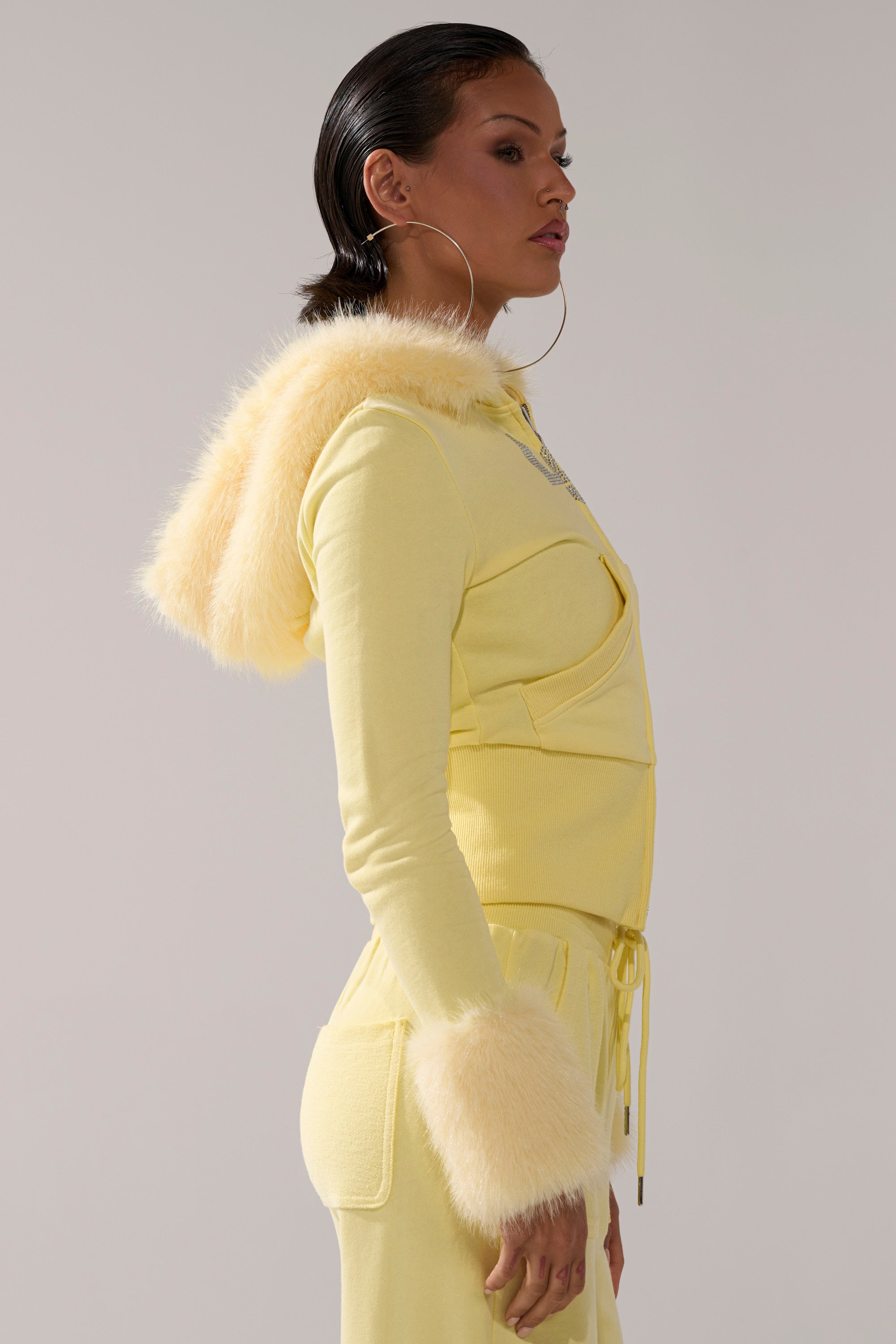 A woman in profile wears the BUTTERFINGER ZIP UP SWEATSHIRT with faux fur trim, matching drawstring pants, slicked-back hair, and large hoop earrings, posing against a neutral background.