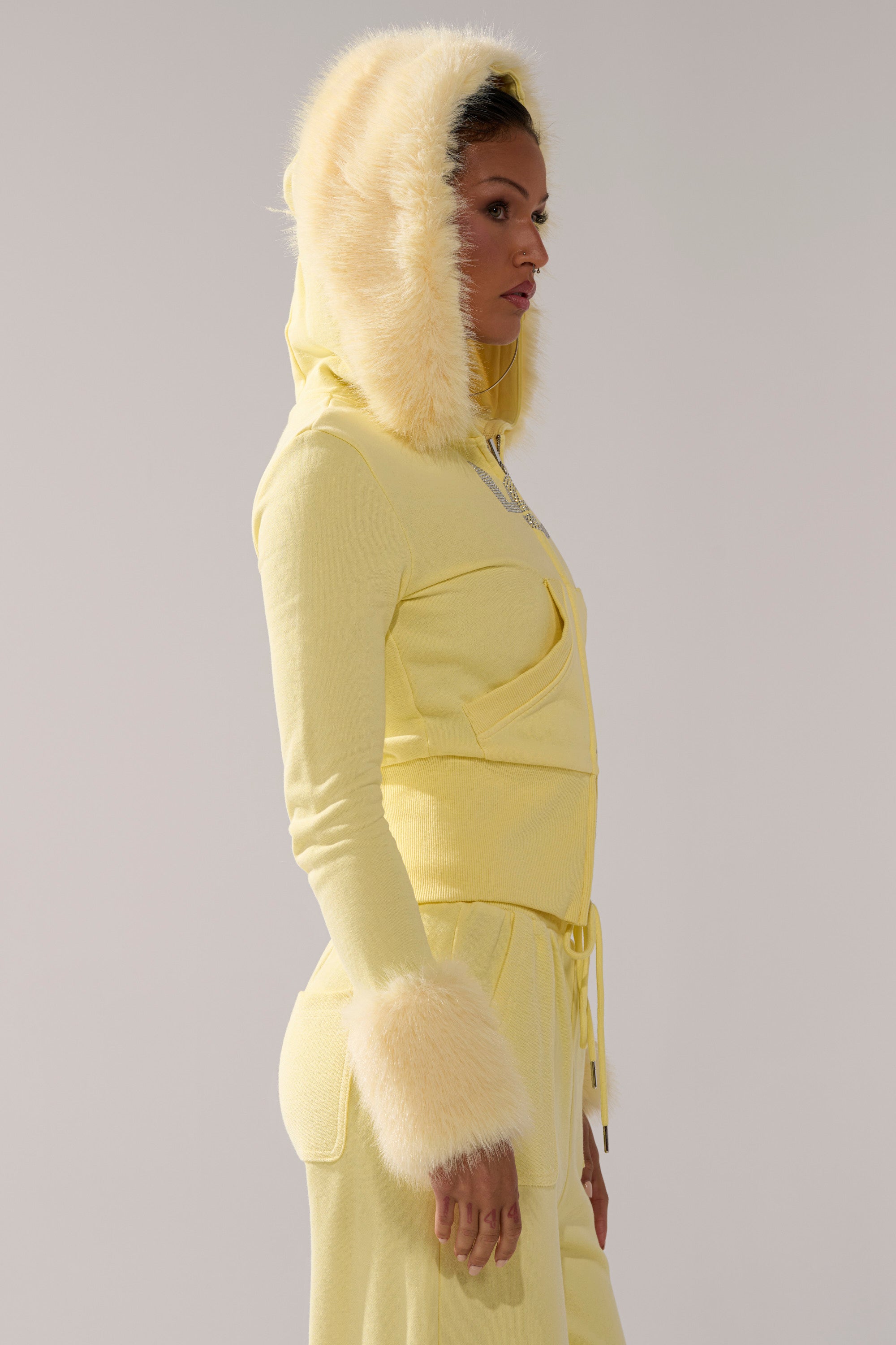 Someone stands in profile against a plain light background, wearing the BUTTERFINGER ZIP UP SWEATSHIRT in pastel yellow, featuring soft faux fur trim on the hood and cuffs for a cozy touch.