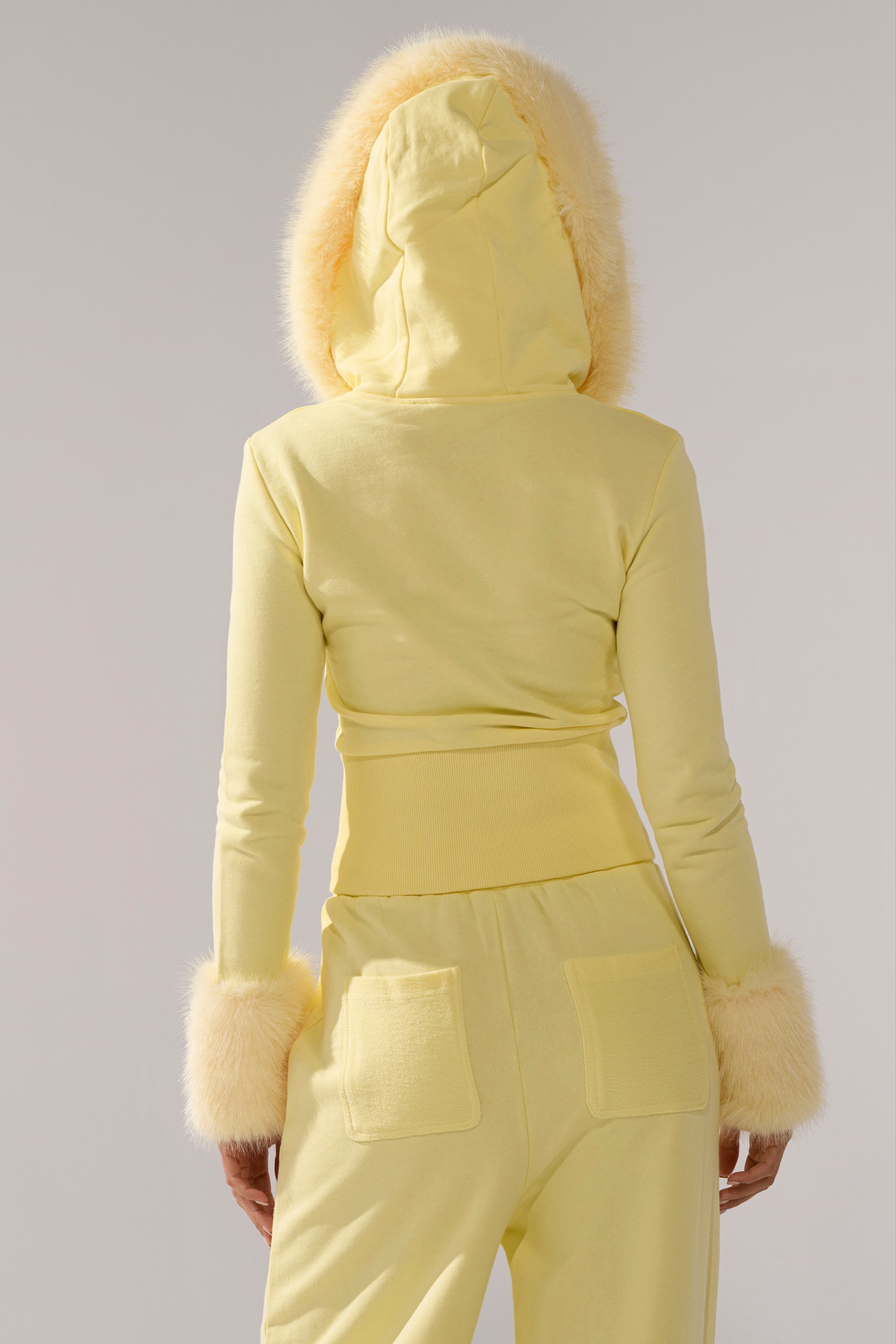 A person is shown from behind wearing the BUTTERFINGER ZIP UP SWEATSHIRT, a pale yellow faux fur trim top, paired with matching yellow pants featuring back pockets, set against a plain background.