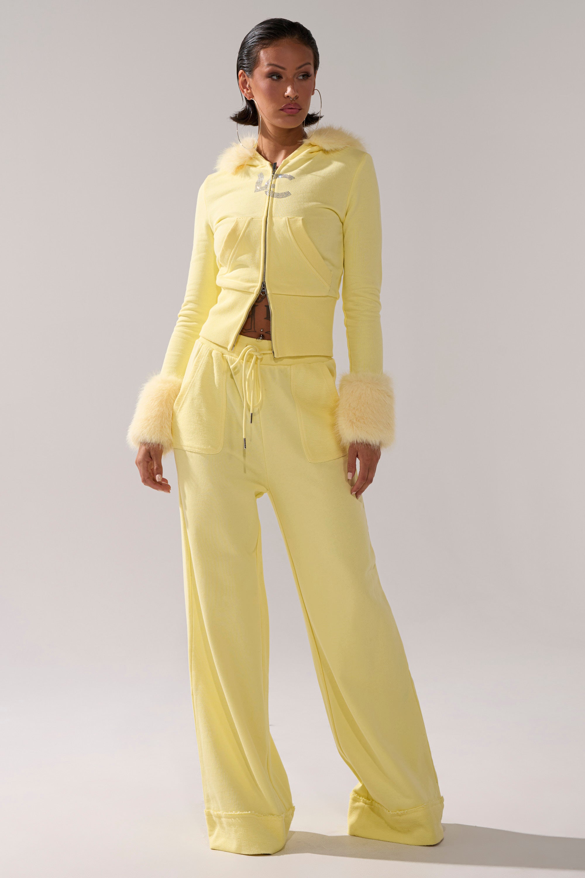 A woman wears the BUTTERFINGER ZIP UP SWEATSHIRT, a pale yellow velour tracksuit with faux fur hood and cuff trim, and matching wide-leg pants. She poses against a simple, light background.