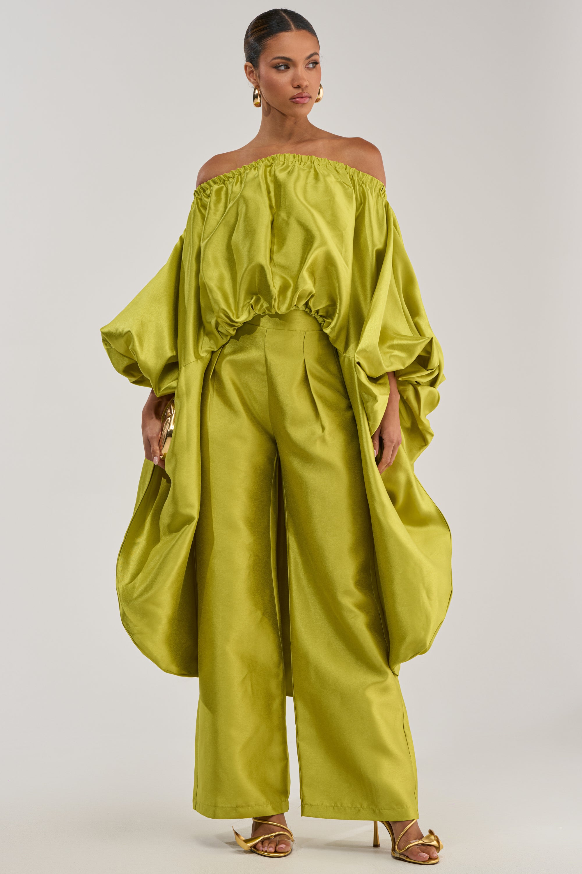 A woman stands against a plain light background in gold sandals, a shiny lime green off-shoulder top with voluminous sleeves and a draped hem, paired with the BY MY SIDE TROUSER IN CHARTREUSE.