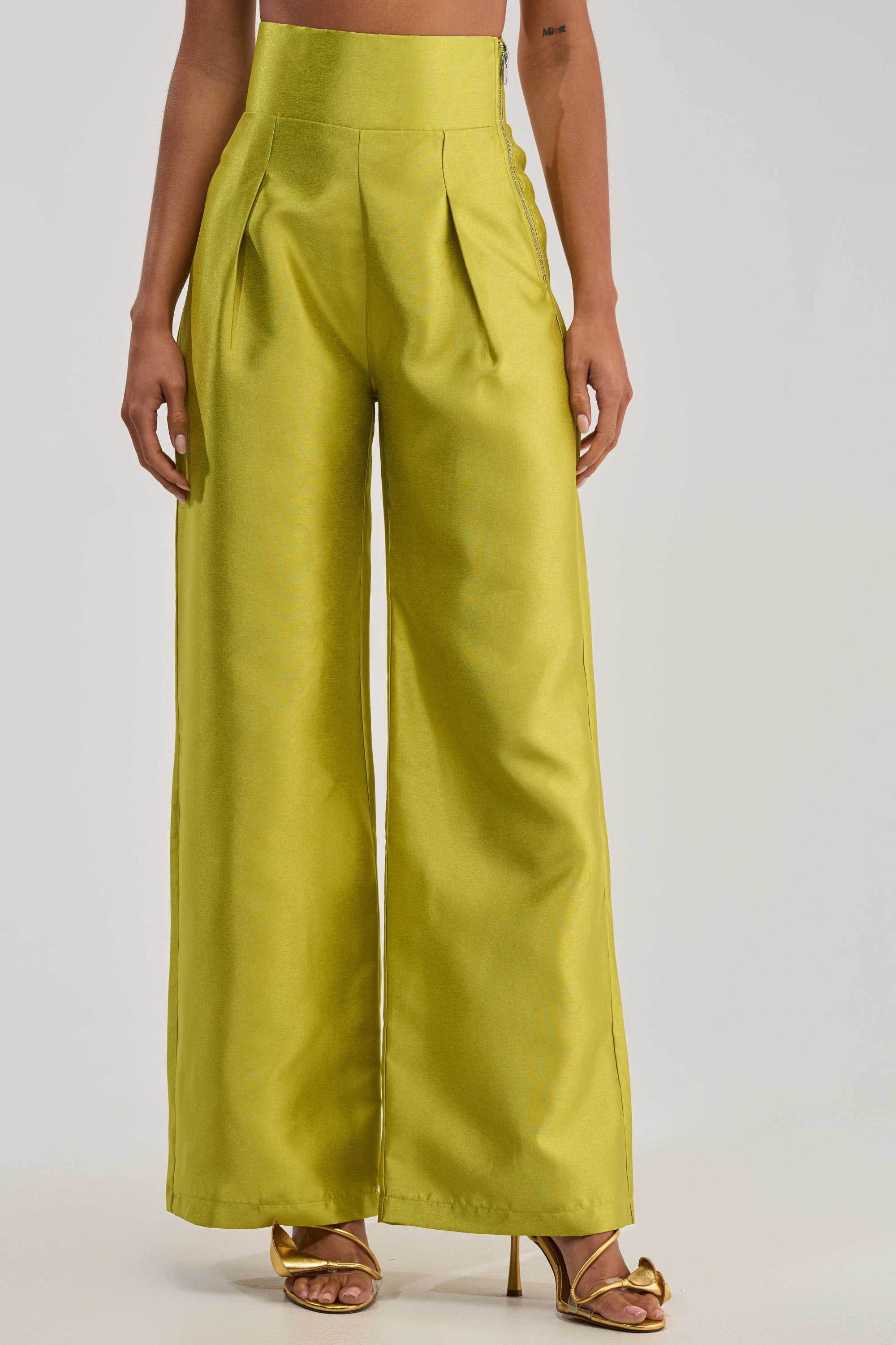 Wearing the BY MY SIDE TROUSER IN CHARTREUSE with a metallic sheen and gold open-toe heels against a light background embodies high-glamour evening style.