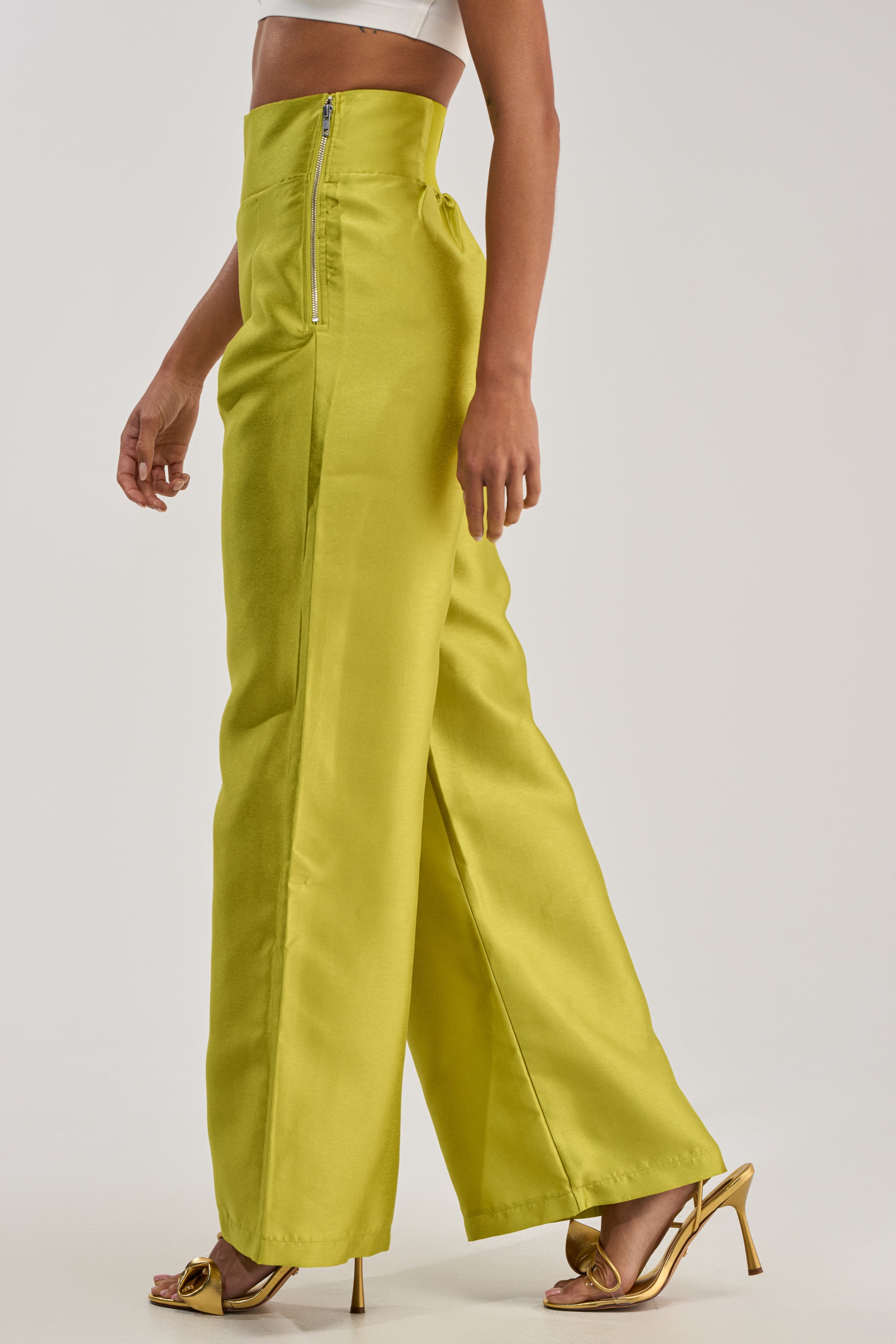 Wearing the BY MY SIDE TROUSER IN CHARTREUSE with a metallic sheen, a white crop top, and gold heels, this person embodies bold high-glamour evening style against a plain light background.