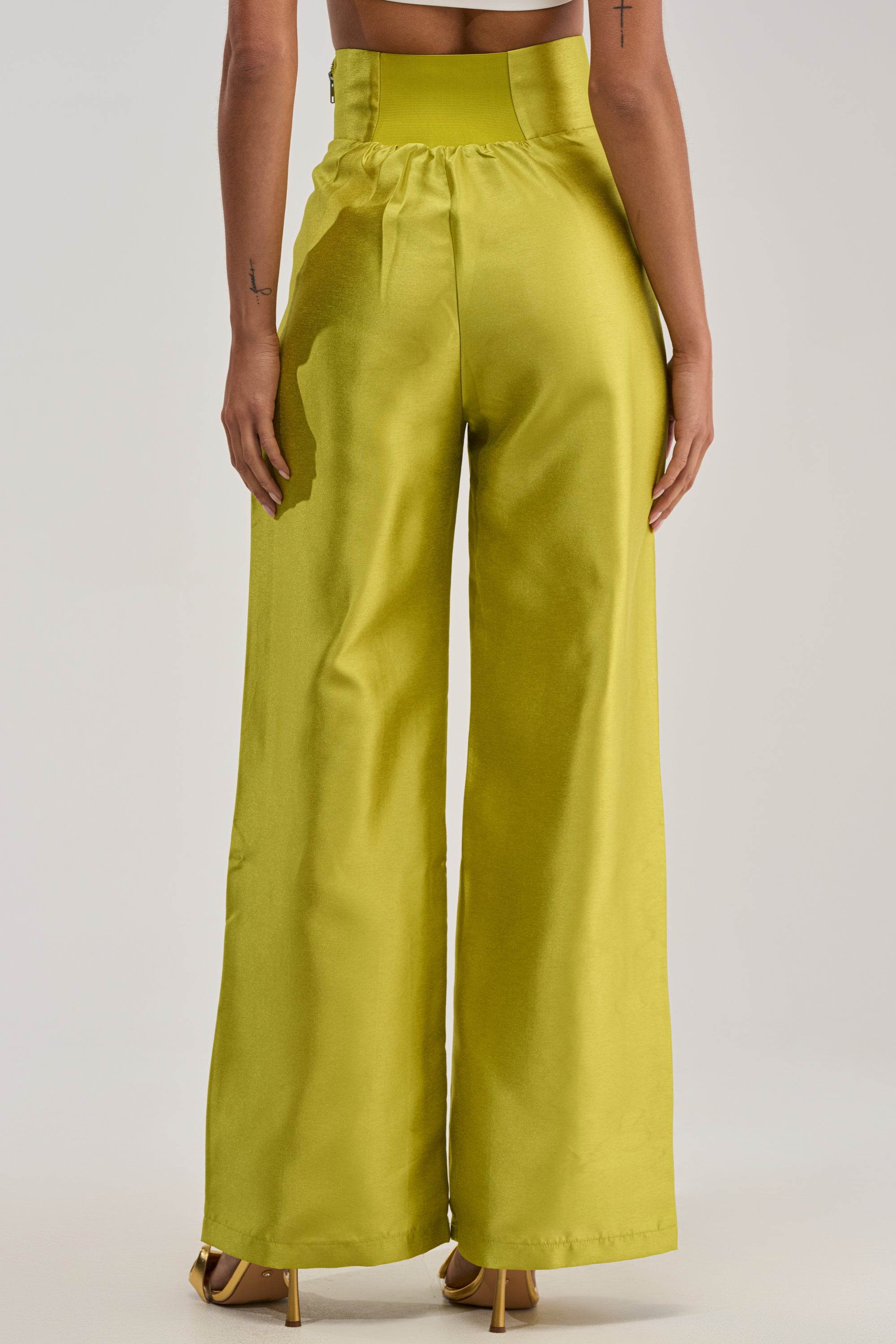 A person stands with their back to the camera wearing the BY MY SIDE TROUSER IN CHARTREUSE—high-waisted, wide-leg satin pants with a shiny metallic finish—styled with high heels for a glamorous evening look.