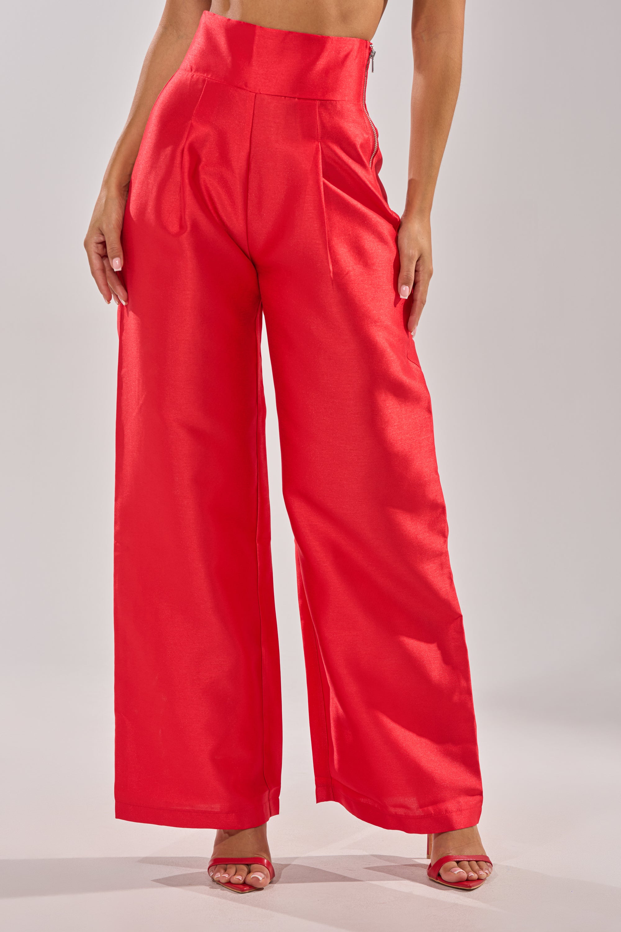 A person wears the BY MY SIDE TROUSER, high-rise wide-leg red satin pants with a shiny finish, paired with matching red open-toe heels. The upper body is cropped out of the image. 0