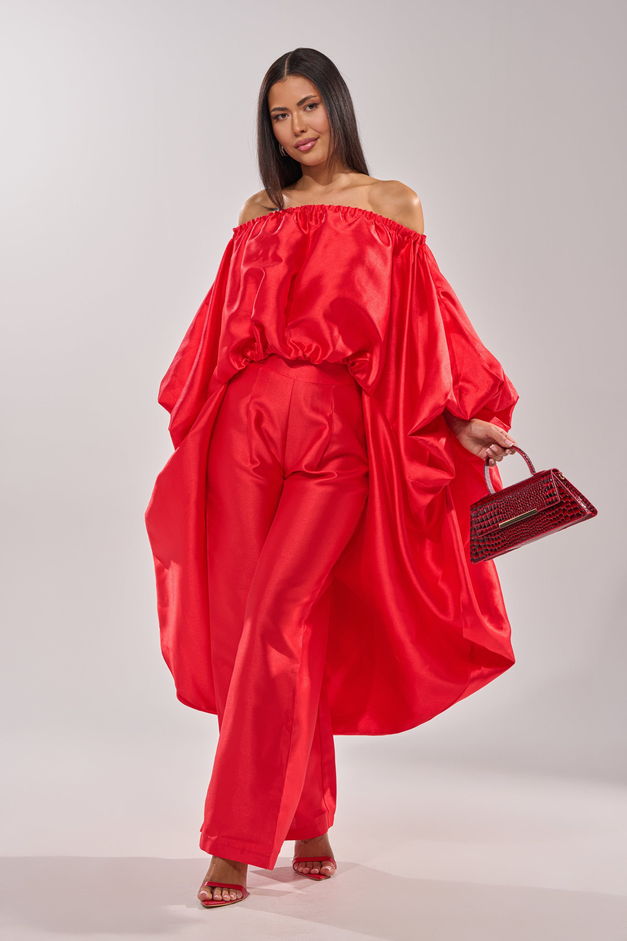 A woman stands against a light background wearing the BY MY SIDE TROUSER, paired with a bright red off-shoulder top, voluminous sleeves, a small textured red handbag, and red open-toe heels. 6