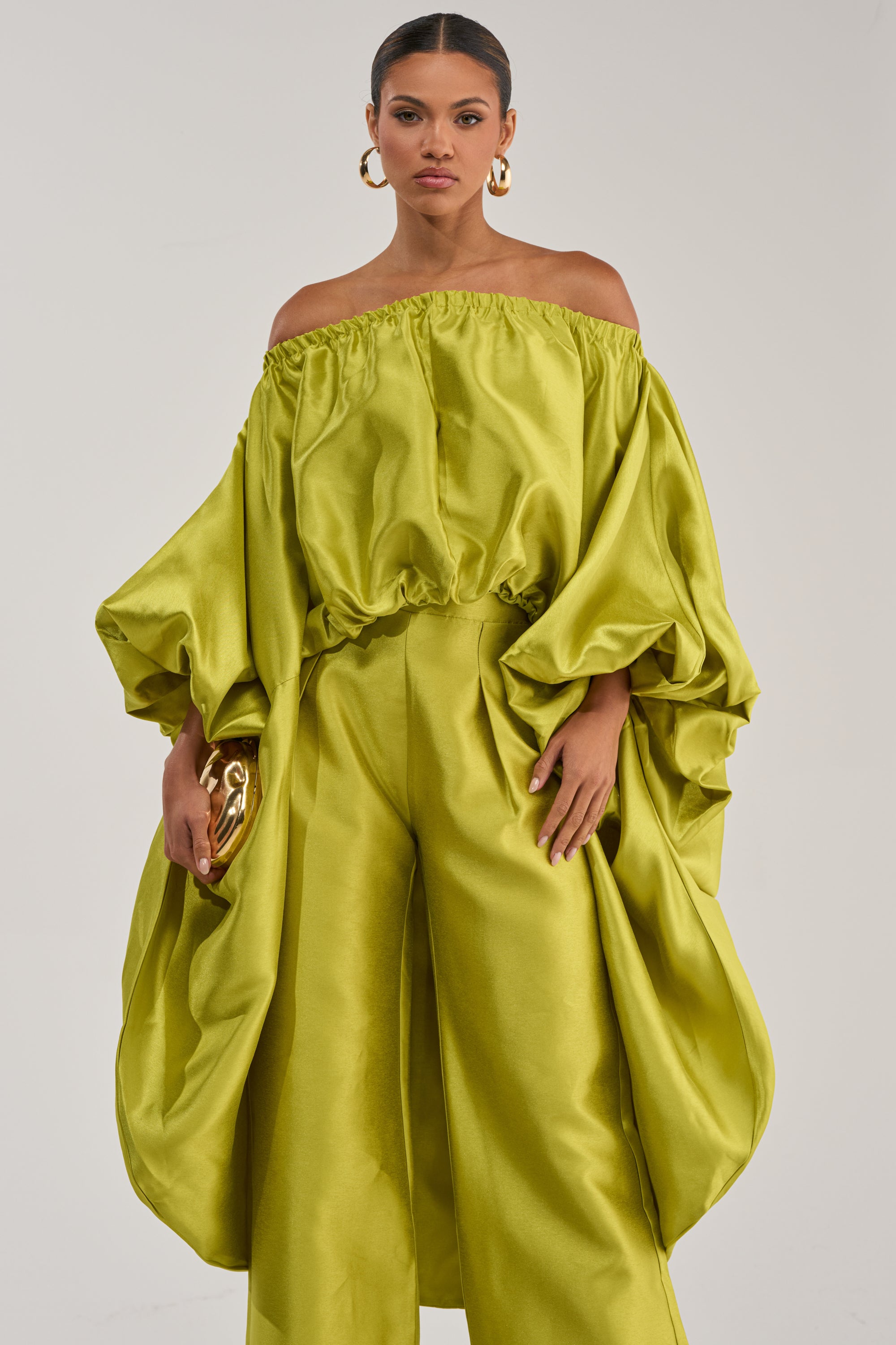 A woman confidently poses against a plain light background wearing the BY MY SIDE HIGH LOW BLOUSE IN CHARTREUSE, styled off-the-shoulder with voluminous sleeves and paired with wide-legged pants. She holds a gold clutch.