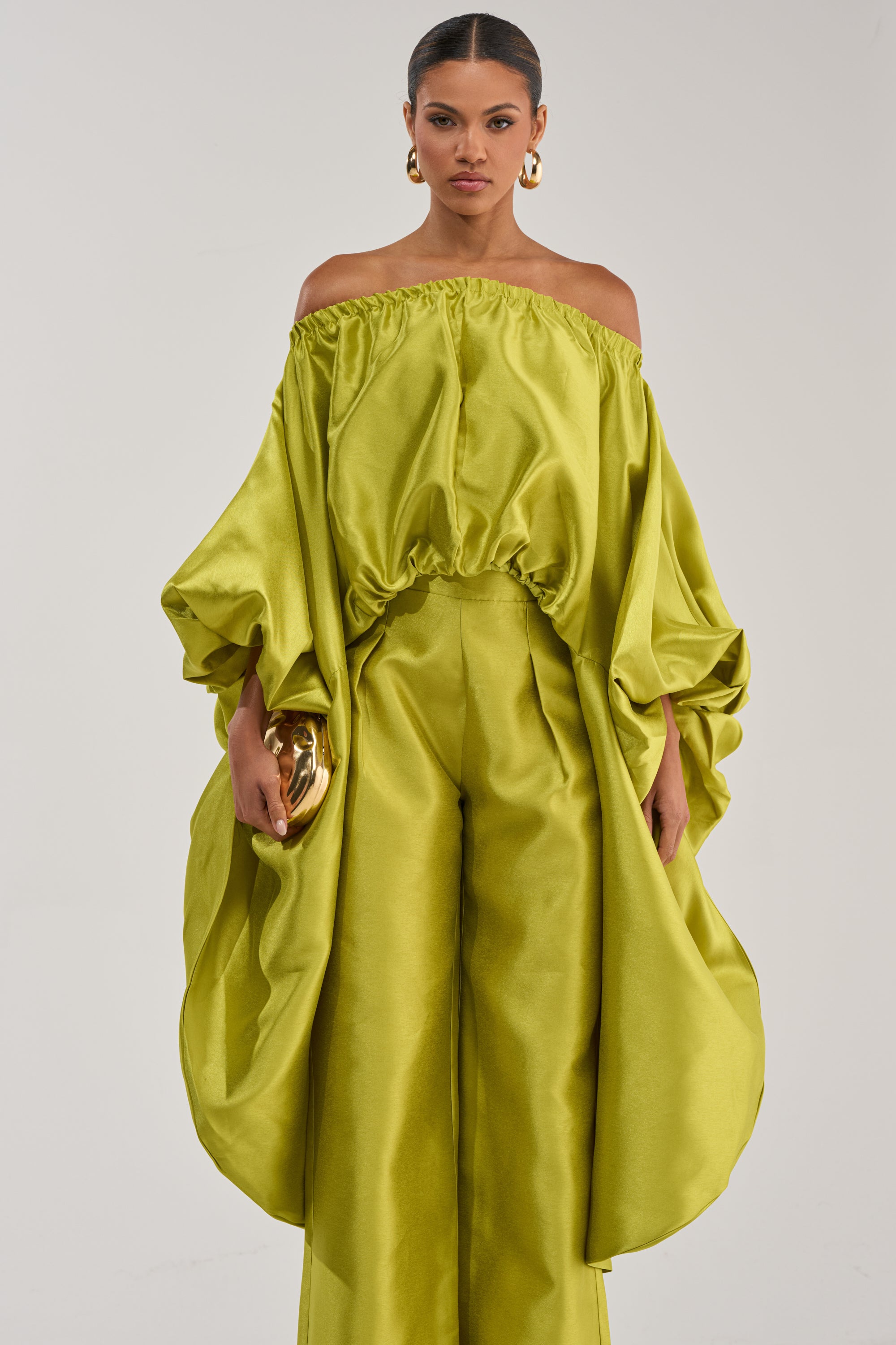 A woman wears the BY MY SIDE HIGH LOW BLOUSE IN CHARTREUSE, an off-the-shoulder, shiny blouse with voluminous sleeves. She pairs it with pants, a small gold clutch, and large gold hoop earrings against a plain background.