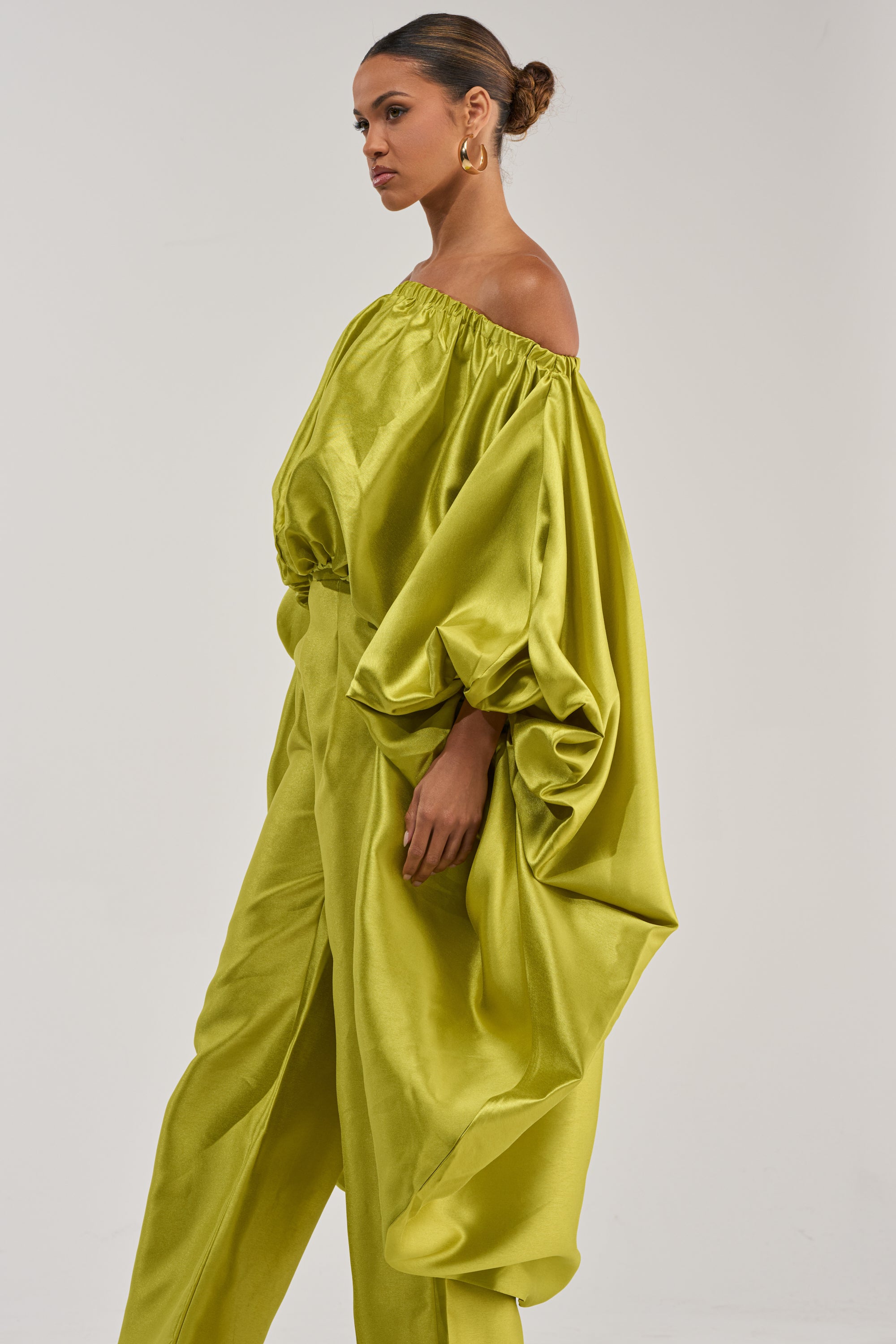 A woman in the BY MY SIDE HIGH LOW BLOUSE IN CHARTREUSE, featuring an off-the-shoulder metallic sheen and billowy sleeves, stands against a white background with matching trousers, gold hoop earrings, and her hair in a bun.
