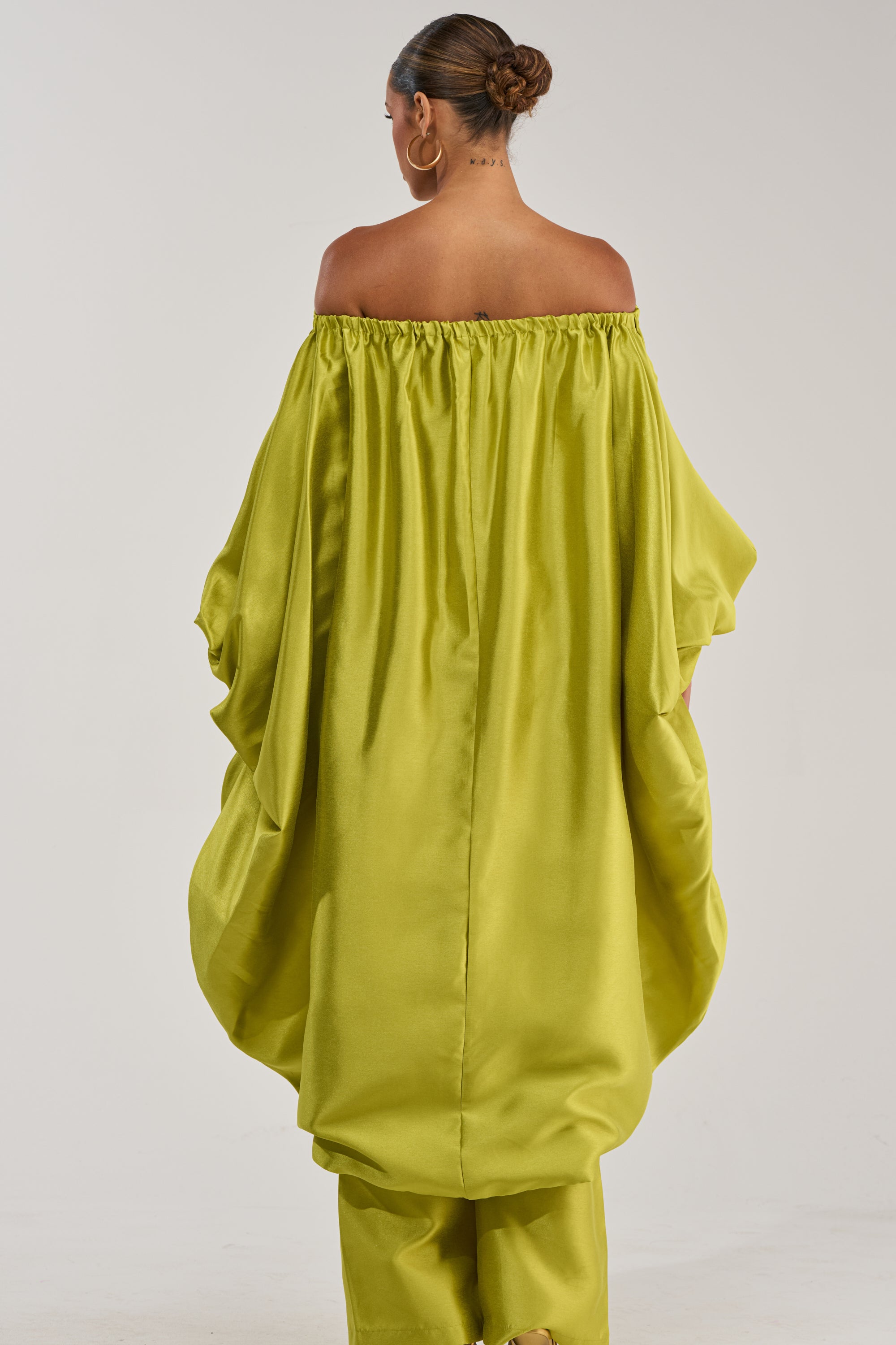 A woman with her back to the camera wears the BY MY SIDE HIGH LOW BLOUSE IN CHARTREUSE, featuring wide sleeves and a metallic sheen, paired with matching pants, a neat bun, and large hoop earrings for a modern formal look.