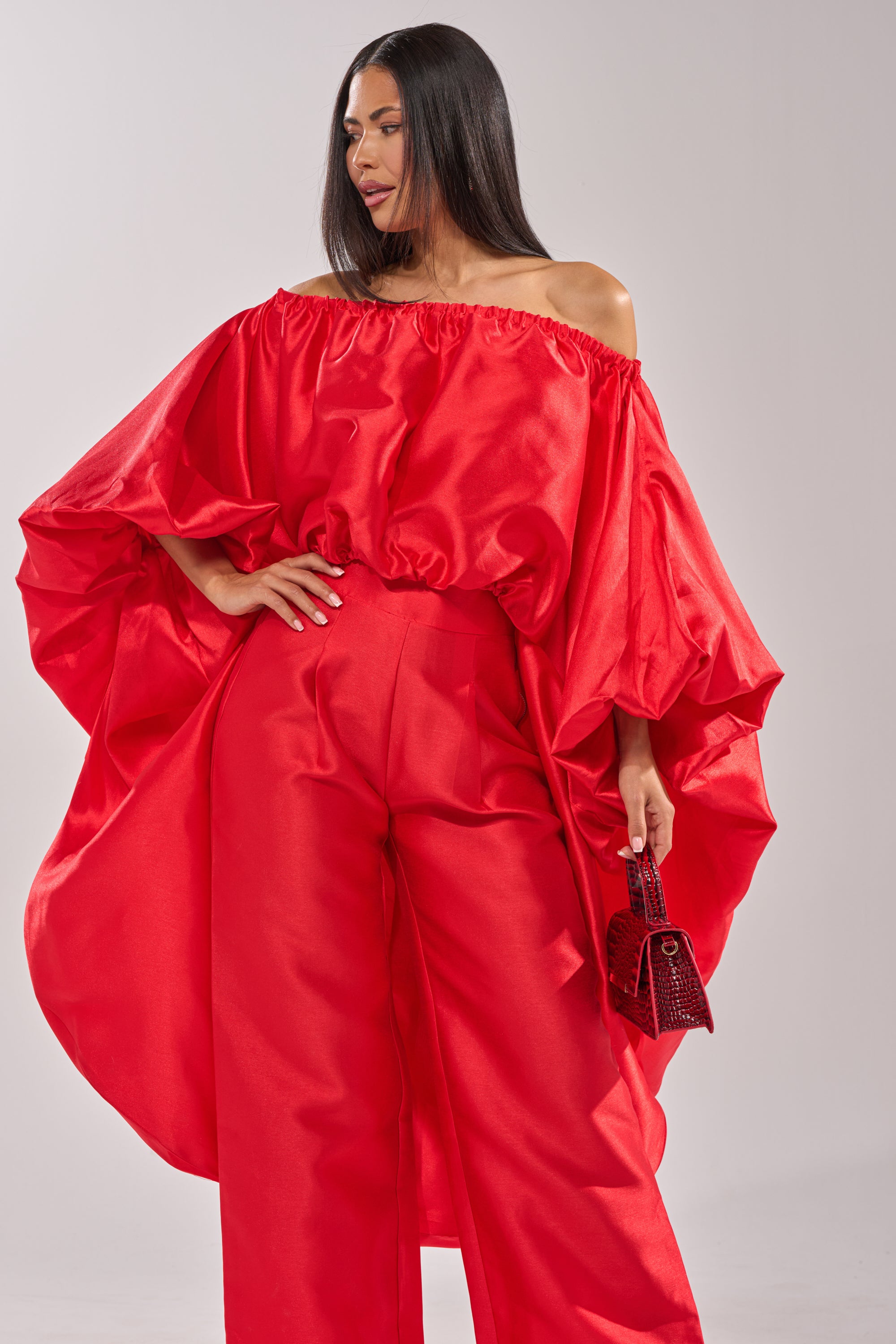 A woman models the BY MY SIDE HIGH LOW BLOUSE in bold red with matching wide-leg pants, holding a small red purse. Her long, straight hair contrasts against a plain light background.