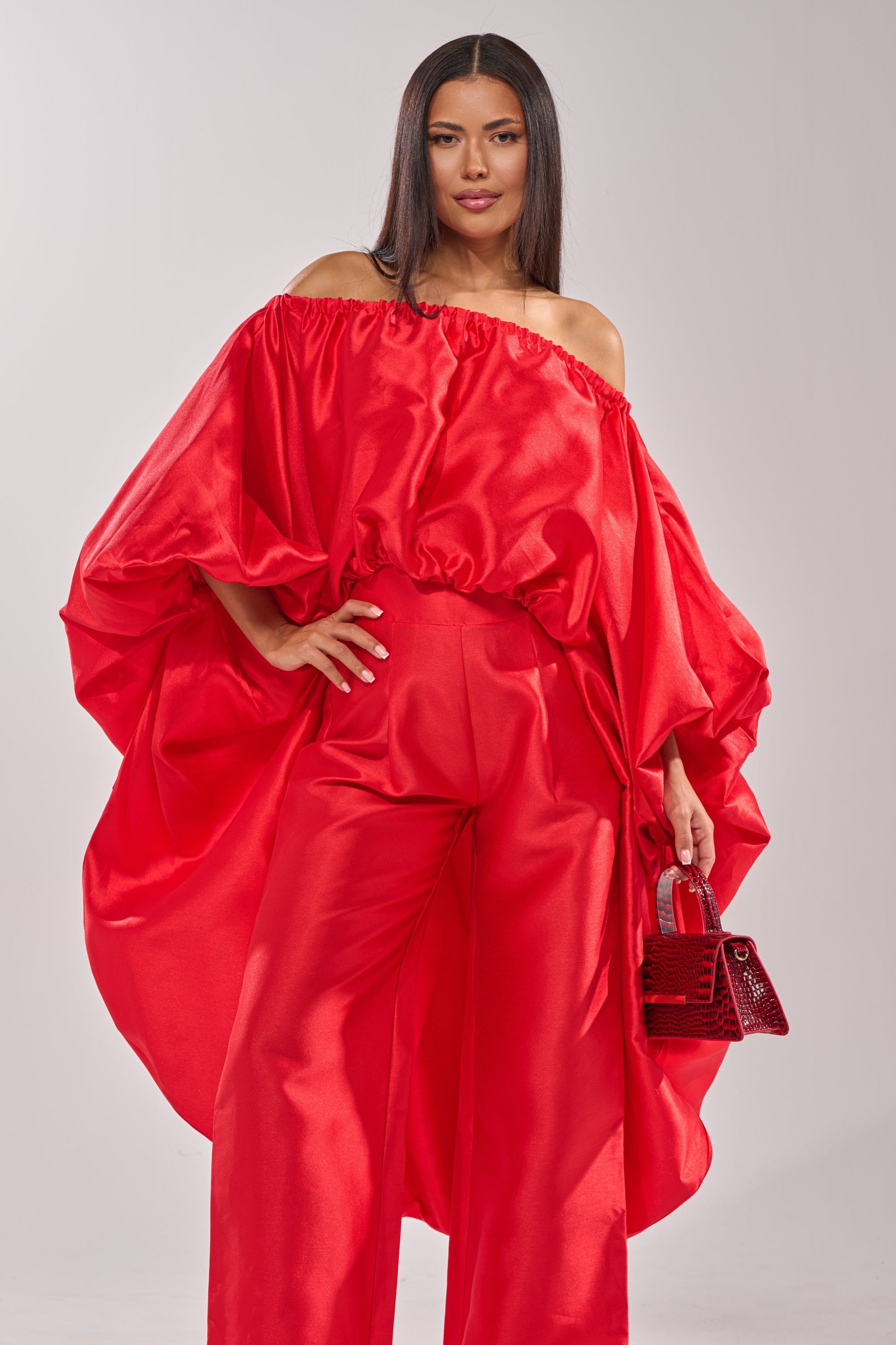 A woman confidently poses in the BY MY SIDE HIGH LOW BLOUSE, a shiny, satin-like off-the-shoulder red top with dramatic sleeves, paired with matching wide-leg pants and a small red handbag. 1