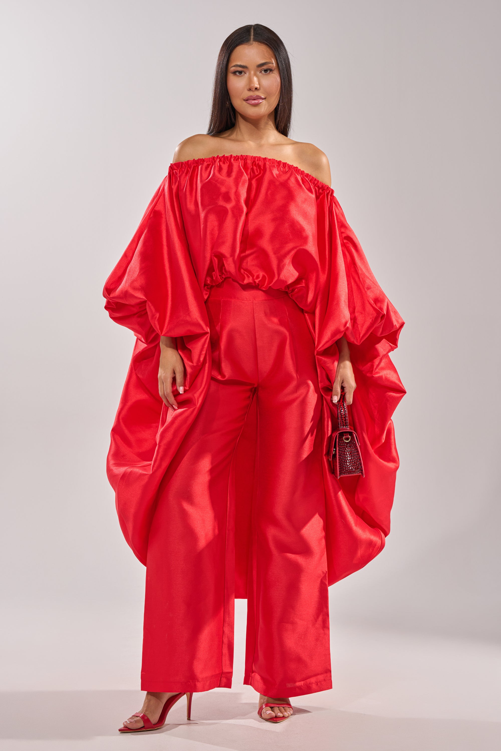 A woman wears the BY MY SIDE HIGH LOW BLOUSE, a bold red, satin-like, strapless top with voluminous sleeves and high-low hem, paired with wide-leg pants, red heels, and a small red handbag against a plain background. 2
