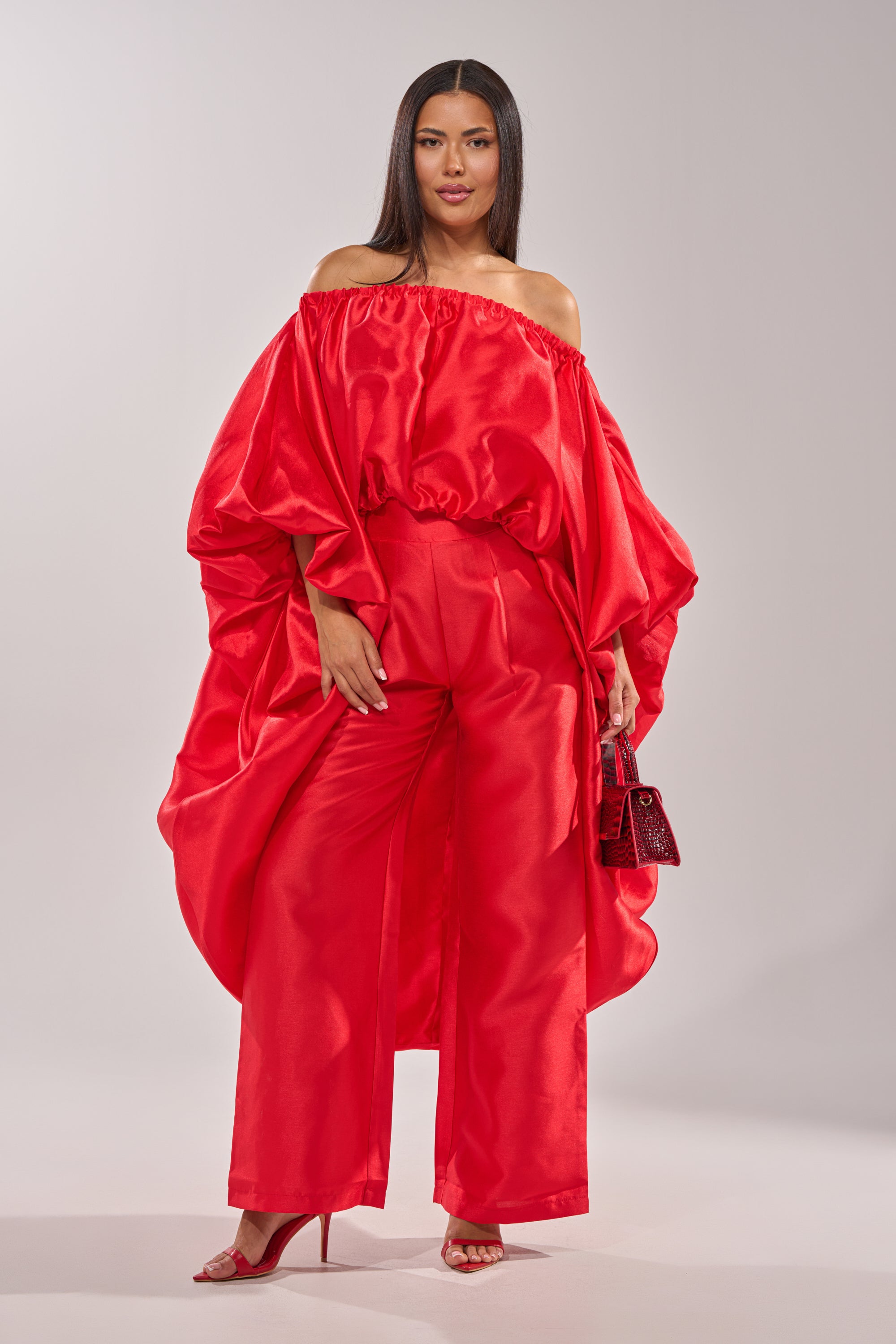 Woman wears the BY MY SIDE HIGH LOW BLOUSE in bold, satin-like red with voluminous sleeves, styled off-the-shoulder with wide-leg pants, red heels, and a small handbag against a simple light gray background. 4
