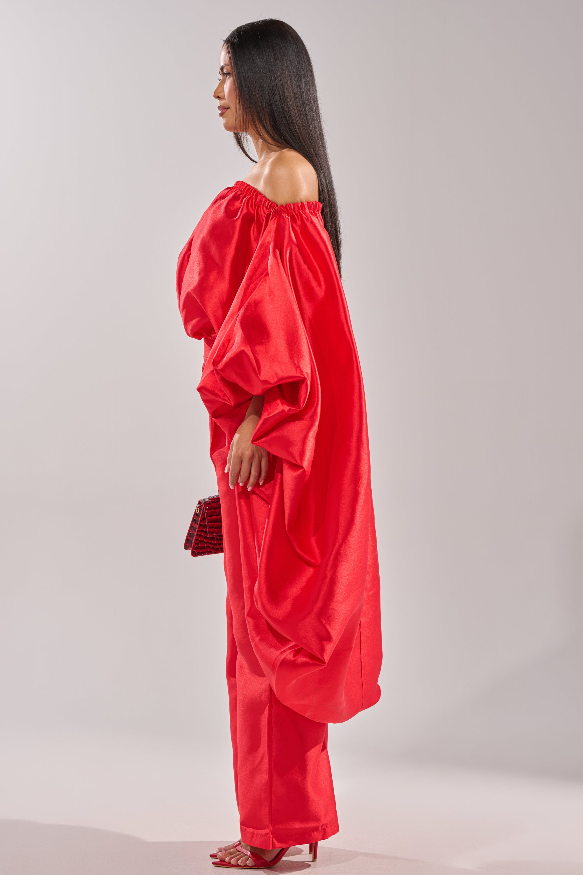 A woman with long, straight dark hair stands in profile against a light background, wearing the BY MY SIDE HIGH LOW BLOUSE in bright red paired with matching pants and heels, and holding a small red bag. 6