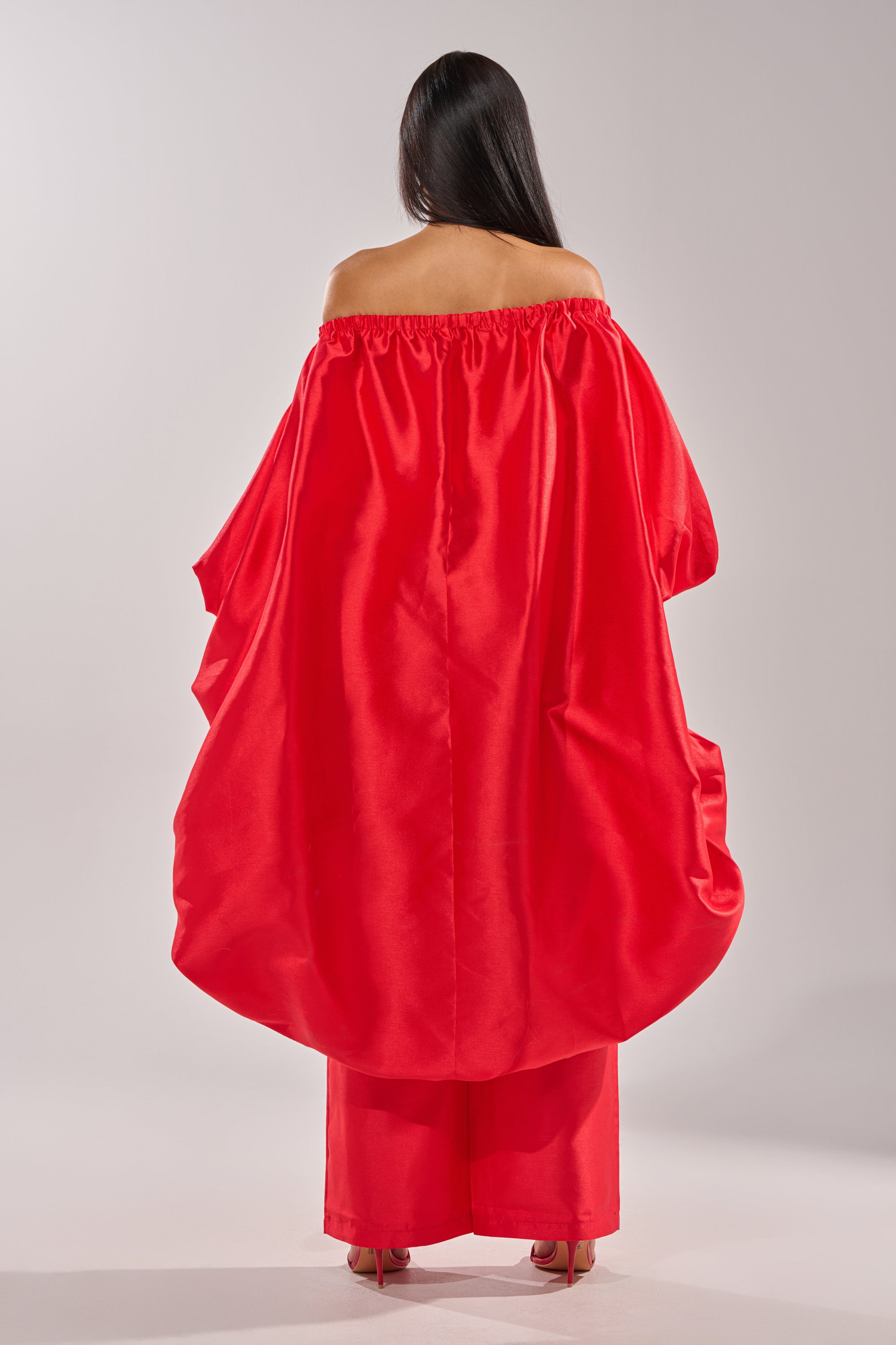 A person with long dark hair faces away, wearing the BY MY SIDE HIGH LOW BLOUSE—an off-the-shoulder, satin-like red top paired with matching wide-leg pants and high heels against a plain light background. 7