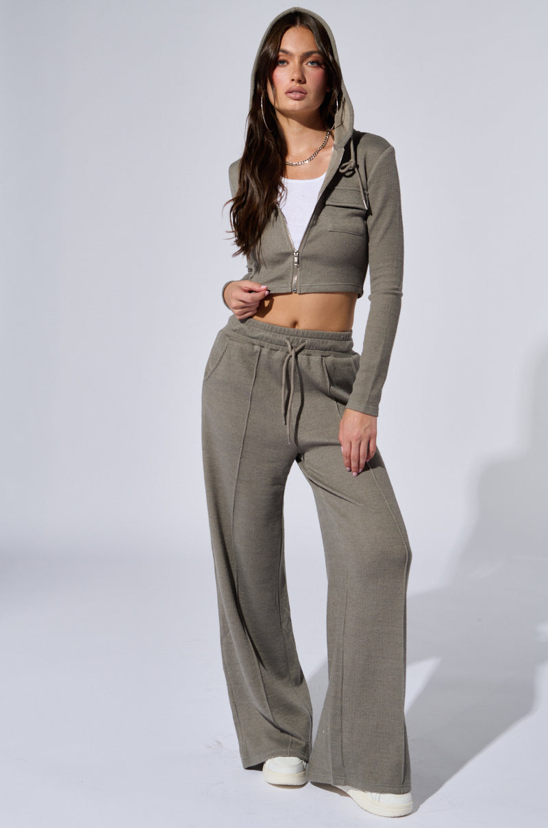 A person poses against a plain background, donning a grey zip-up cropped hoodie over a white shirt and the "EYES ON ME WIDE LEG SWEAT PANT IN GREY," which features a drawstring waist. The high-rise design is complemented by white sneakers and long, wavy hair. The person has a casual, confident stance. 0