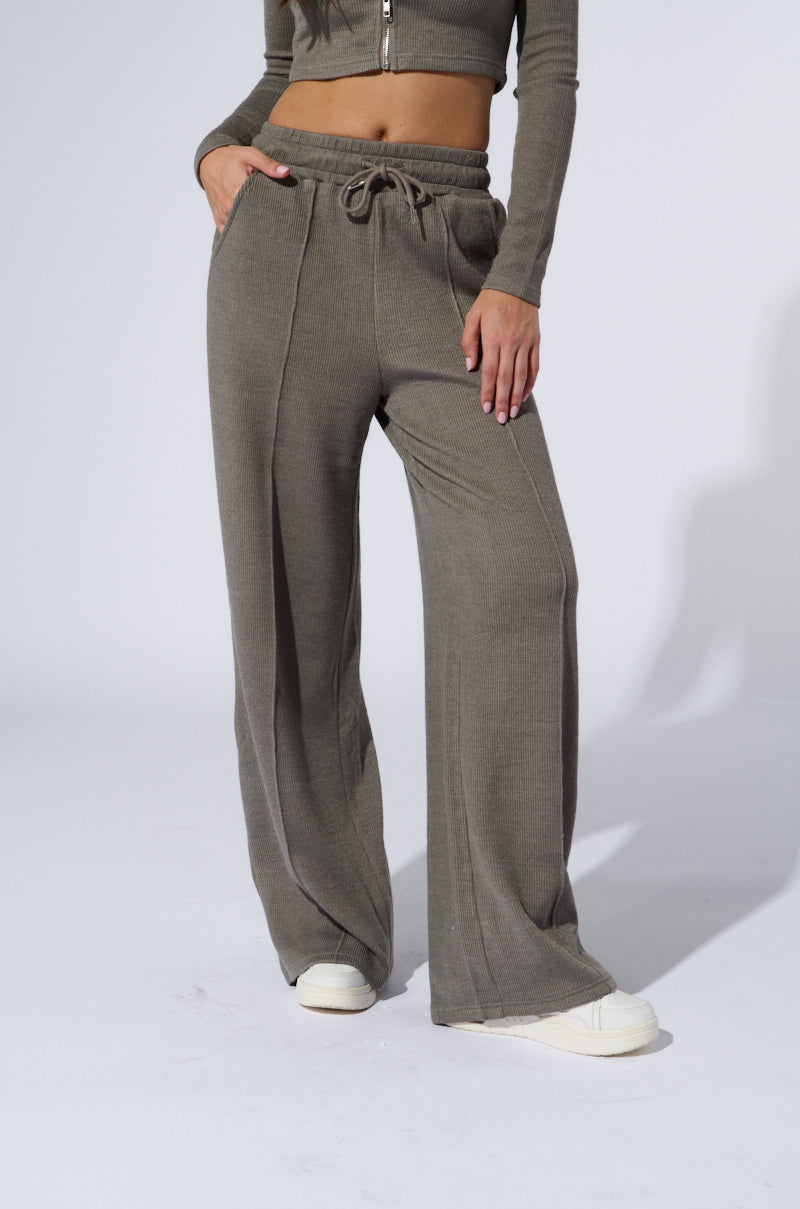 A woman stands against a plain background, wearing a long-sleeve, zip-up cropped top and matching "Eyes on Me" high-waisted wide-leg sweat pants in grey. She has one hand in a pocket of her pants, and she is wearing white shoes. The stretchy ribbed knit fabric of the outfit offers both comfort and style. 1