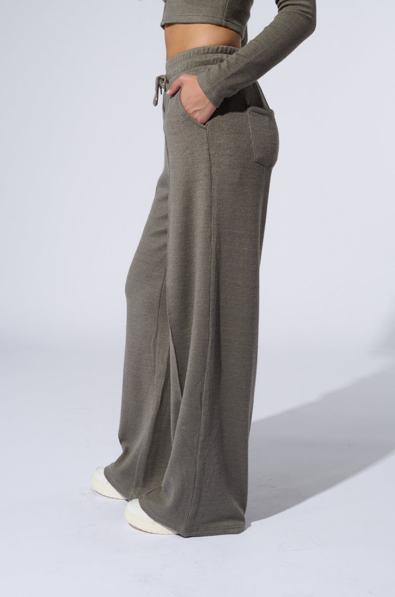 A person stands facing slightly to the left, wearing high-rise, wide-leg sweat pants in grey. The stretchy ribbed knit pants have a drawstring waist and pockets, complemented by a matching long-sleeve top. White sneakers are partially visible. 2