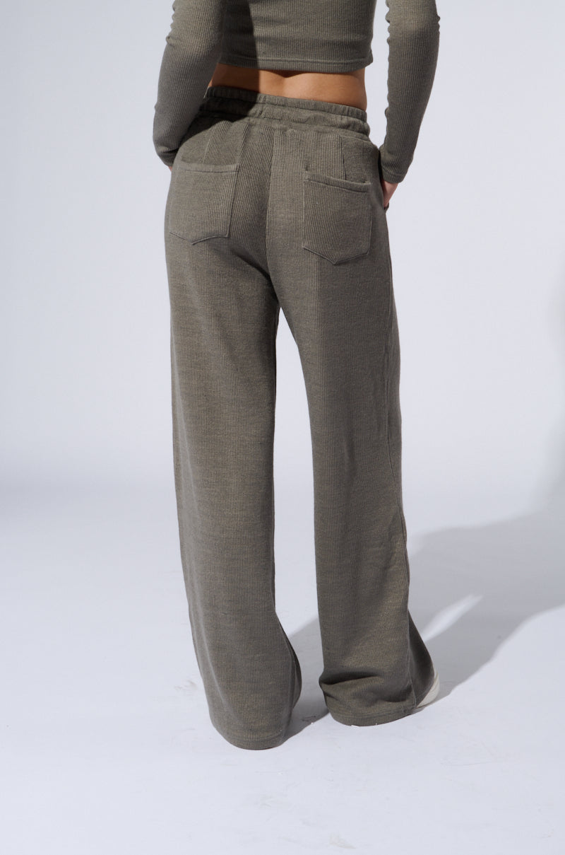 A person is shown from the back, modeling grey wide leg sweat pants. The pants have two back pockets and a loose, relaxed fit, accentuated by their stretchy ribbed knit texture. The individual is also wearing a matching grey knit top that exposes the midriff. The background is plain, light grey. 3