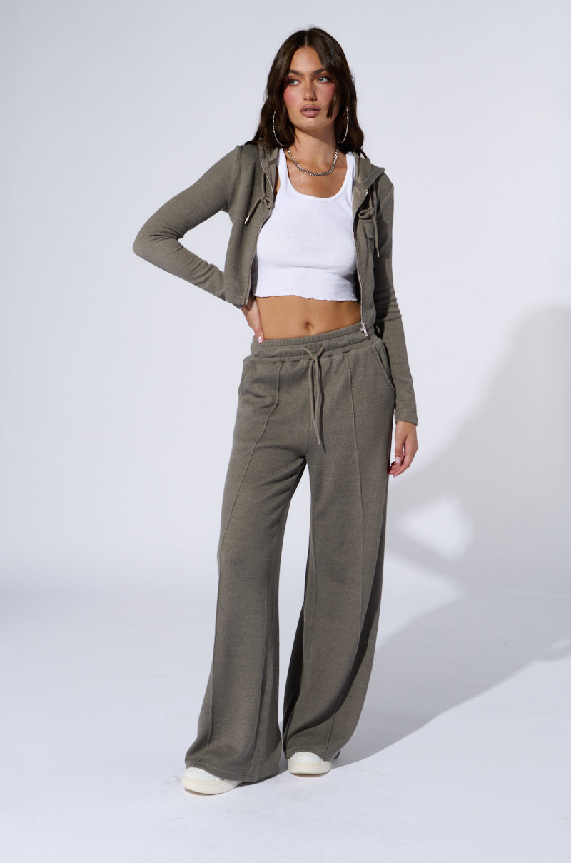 A person with long dark hair stands against a plain background, wearing a matching grey loungewear set featuring the "EYES ON ME WIDE LEG SWEAT PANT IN GREY" and a zip-up hoodie. They have paired it with a white cropped tank top, white sneakers, large hoop earrings, and necklaces that highlight the high-rise design of the sweatpants. 5
