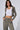 A woman with long dark hair stands with her hands in her pockets, wearing a white crop top and the EYES ON ME RIB KNIT ZIP UP IN GREY along with matching drawstring pants. She accessorizes with hoop earrings and a silver necklace against a plain, light grey background.