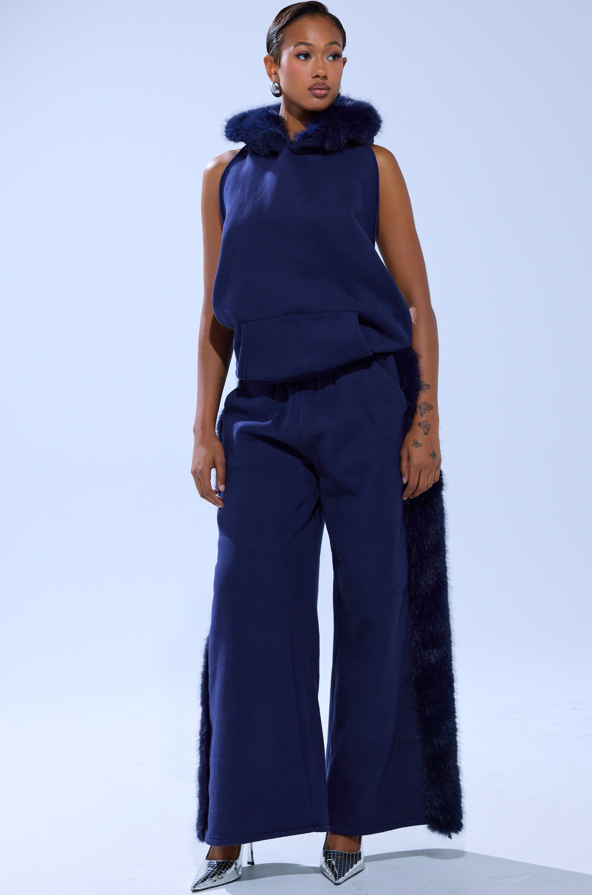 A woman poses in the LUXE FUR JOGGER IN NAVY, a cotton-blend sleeveless top and matching wide-leg pants, both detailed with faux fur trim. She completes the stylish look with pointed silver heels against a plain light background.