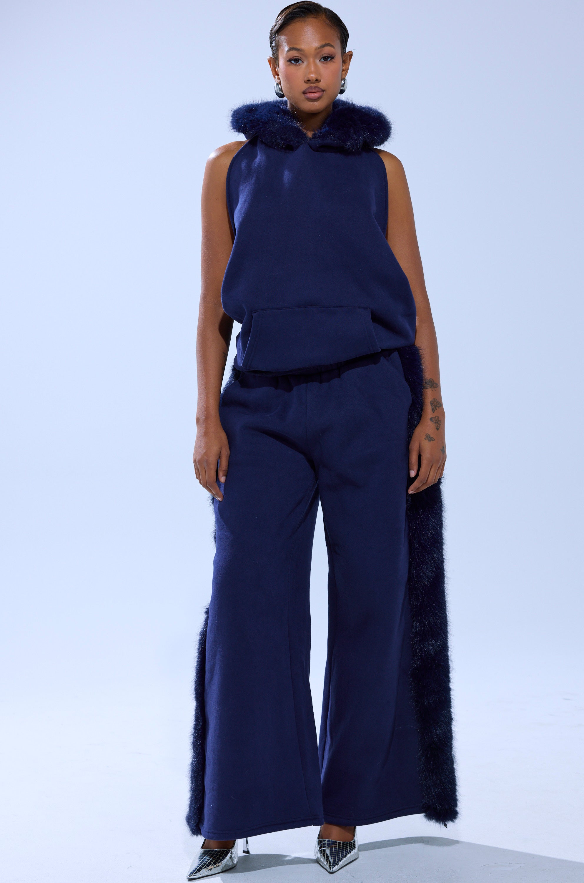 A woman stands facing forward in the LUXE FUR JOGGER IN NAVY, a sleeveless hooded top with faux fur trim and matching wide-leg joggers with fur-accented sides. She pairs it with pointed silver heels and wears her hair slicked back.