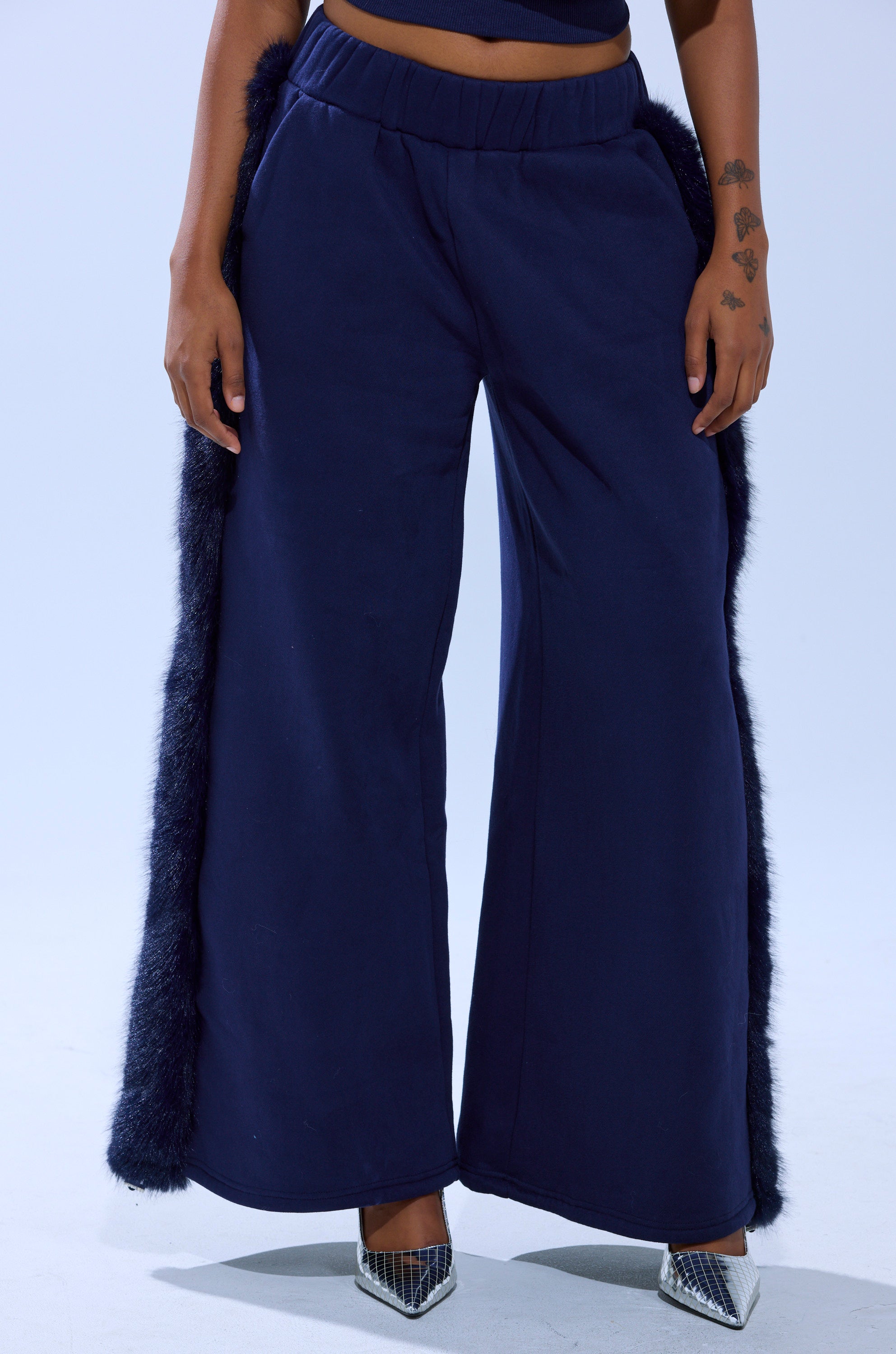 A person models the LUXE FUR JOGGER IN NAVY, featuring black faux fur trim and silver pointed-toe heels, standing against a light background with only the lower half visible.