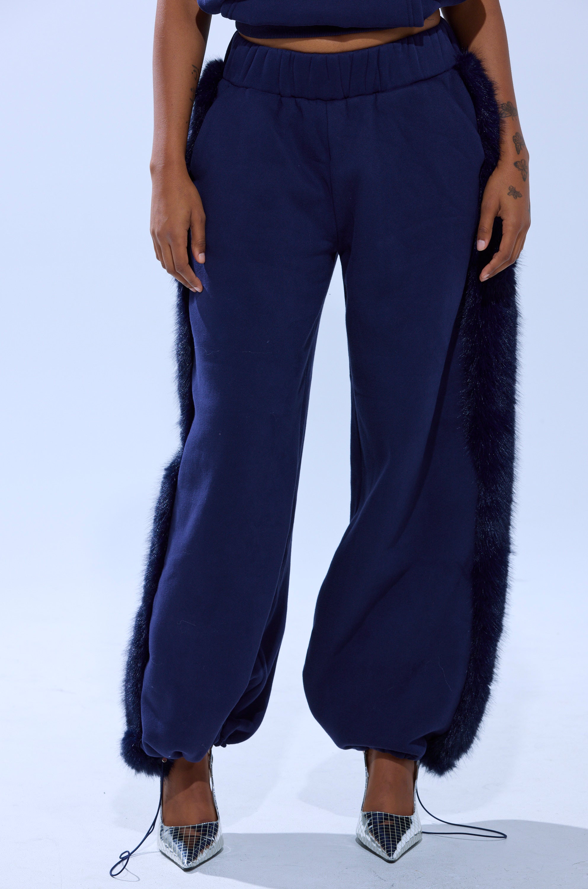 Wearing the LUXE FUR JOGGER IN NAVY with faux fur trim and silver pointed-toe heels, the photo captures the look from shoulders to feet against a plain light background.