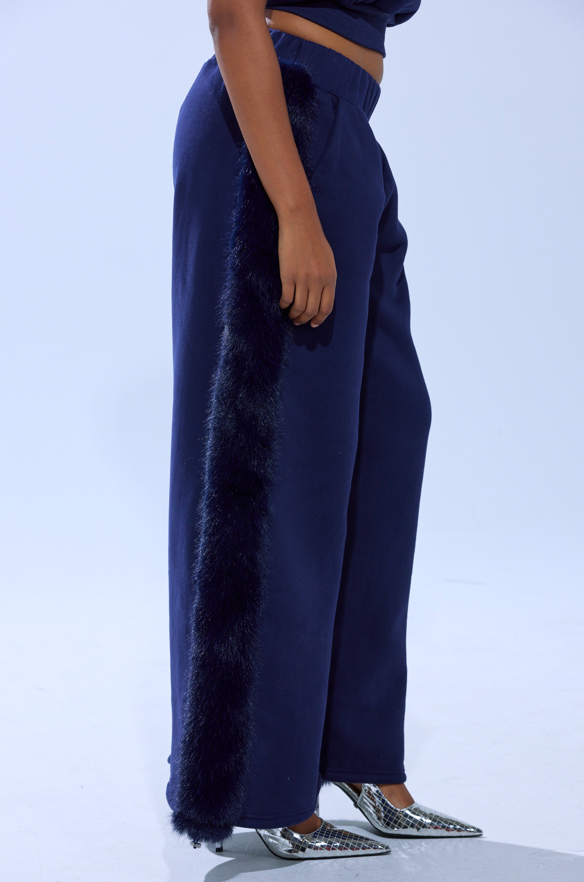 Wearing the LUXE FUR JOGGER IN NAVY with a faux fur side stripe, paired with a matching navy top and silver pointed-toe heels, someone stands against a plain light background.