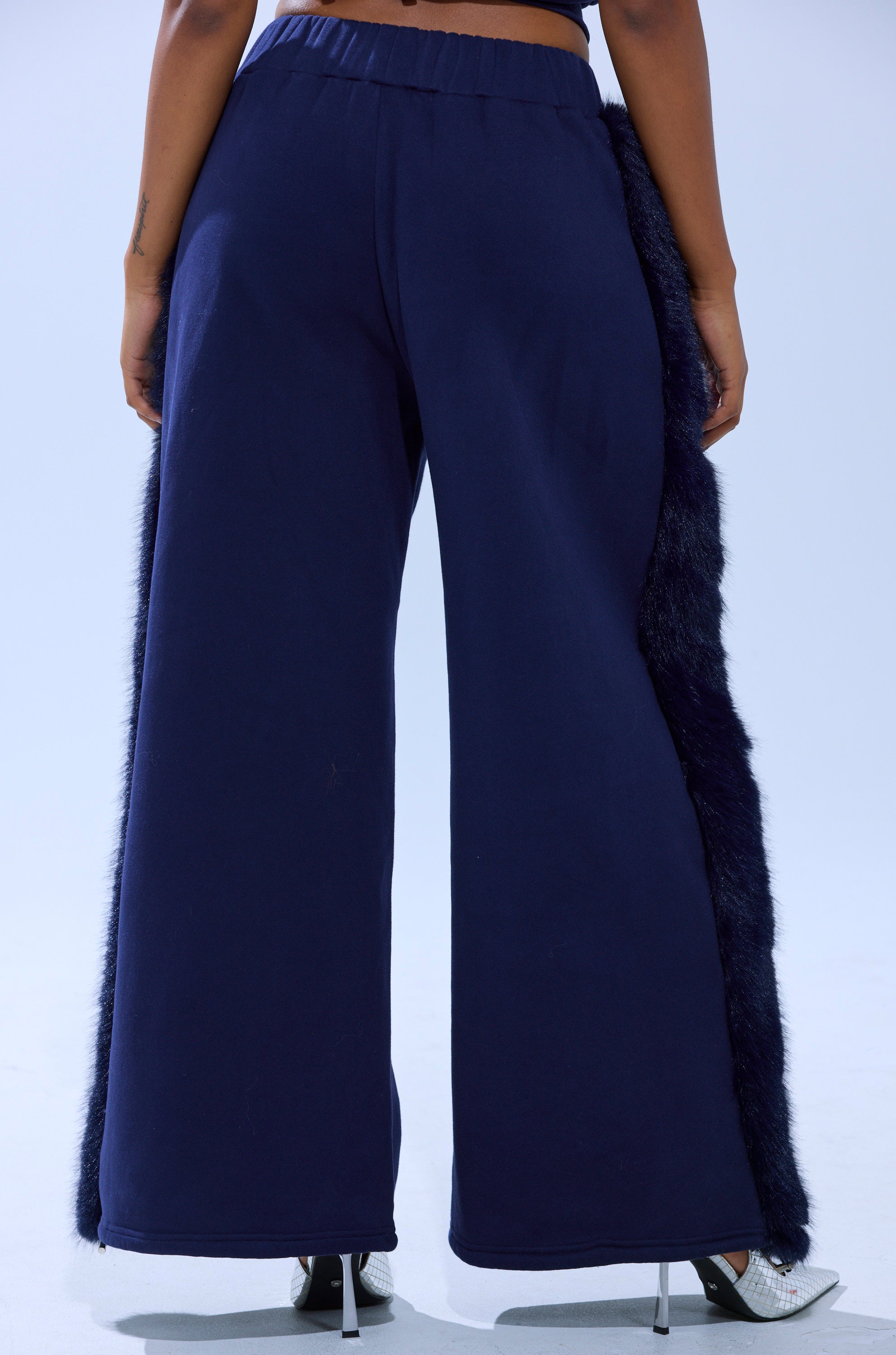 Someone is shown from the back wearing the LUXE FUR JOGGER IN NAVY, featuring faux fur trim on the sides, paired with silver high-heeled shoes.