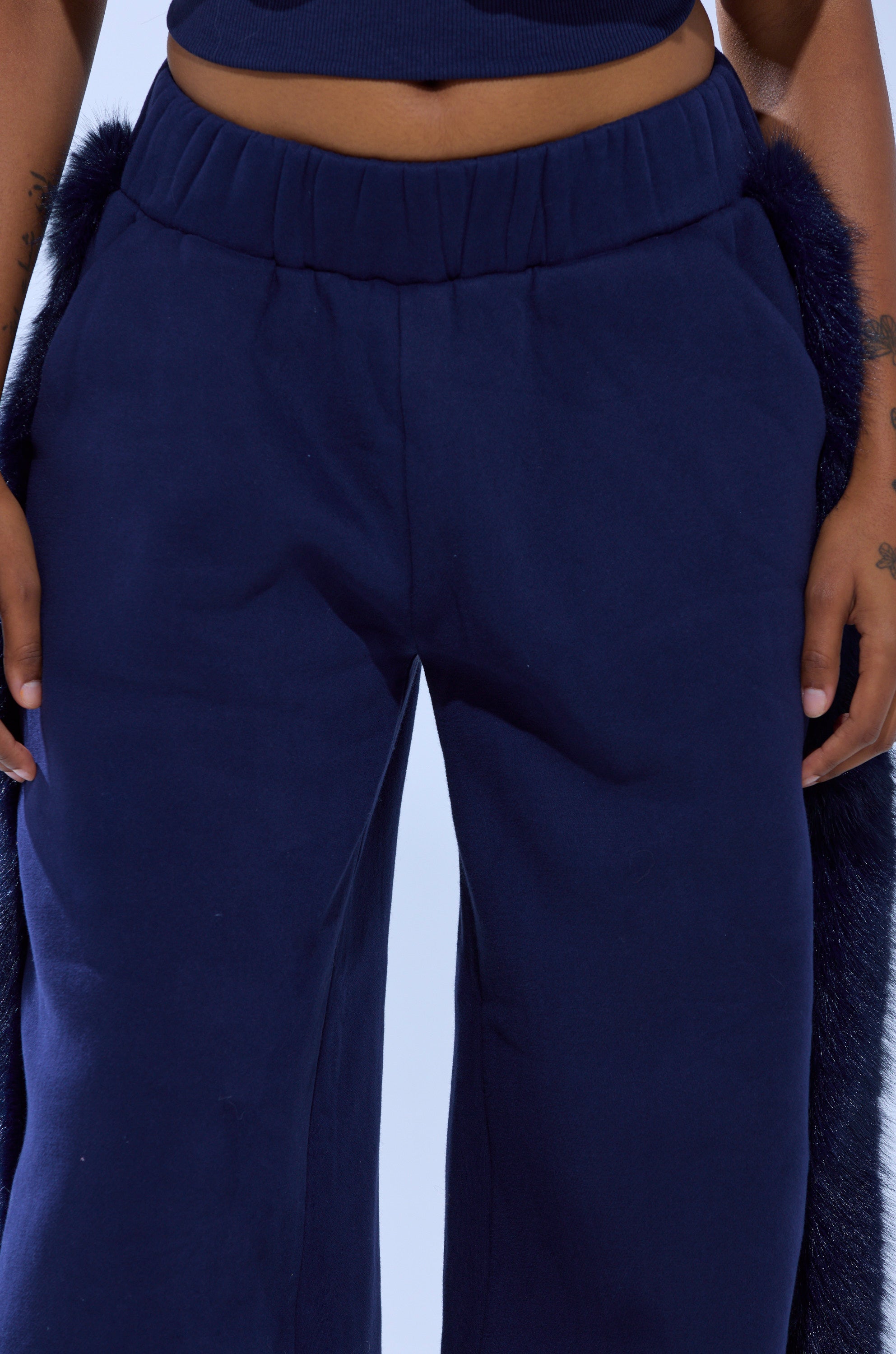 A person wearing LUXE FUR JOGGER IN NAVY—high-waisted, dark blue wide-leg pants with faux fur trim—paired with a matching blue crop top, standing with arms at their sides.